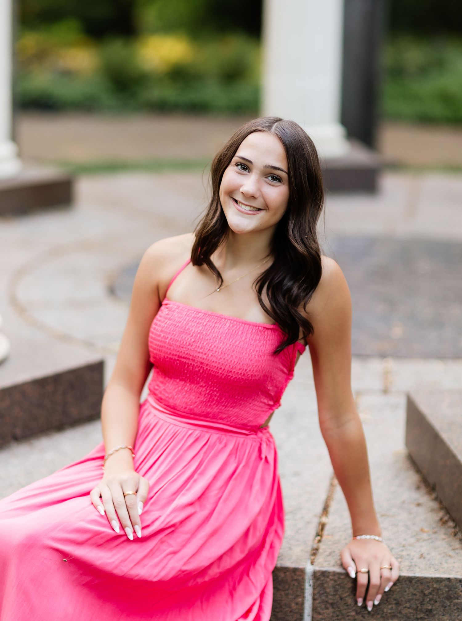 Smiling young woman in hot pink dress sitting on stone steps in an outdoor garden setting.