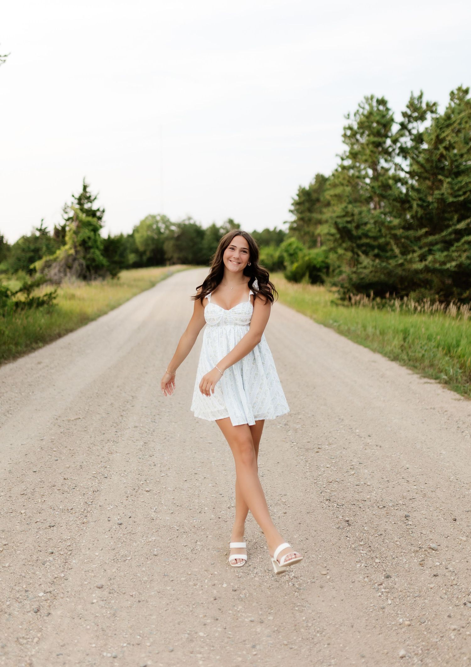 Smiling girl in light blue dress walking on a rural gravel road lined with green trees.