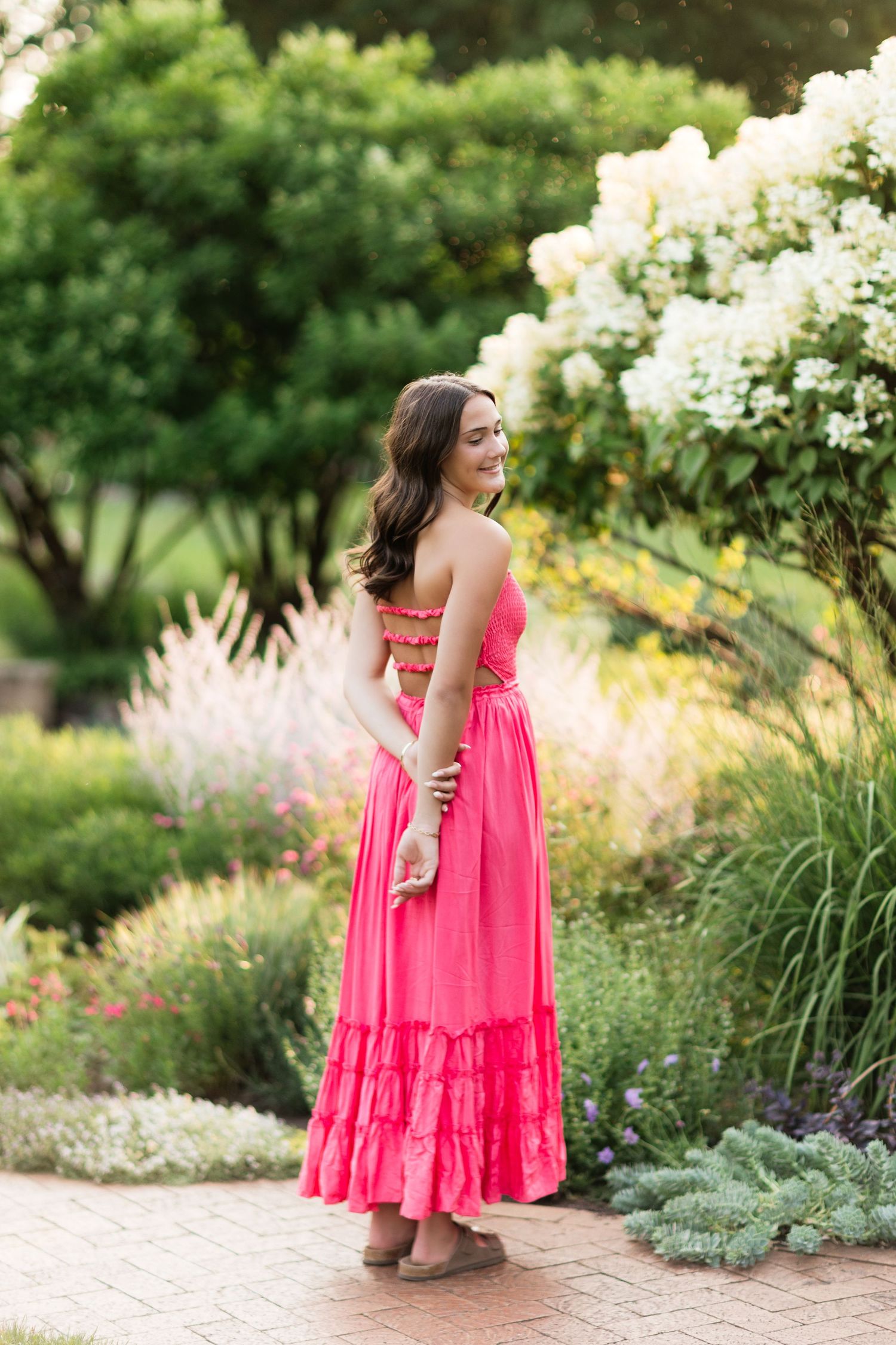 Brunette woman in coral maxi dress posing from behind in a colorful garden.
