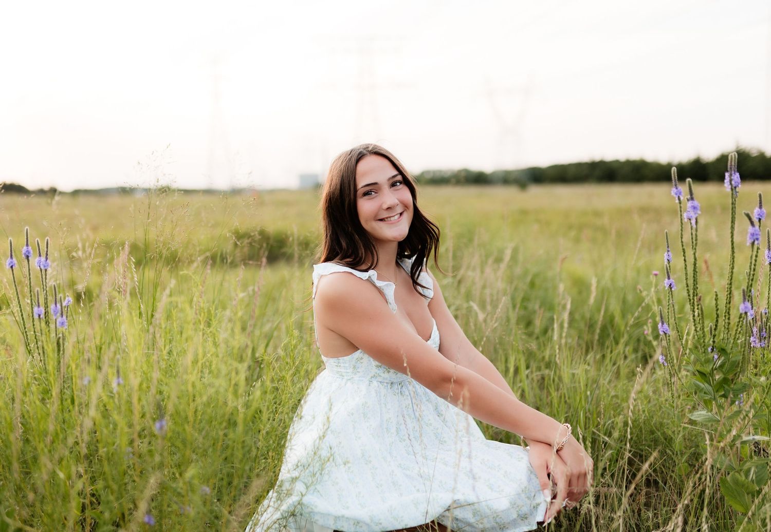 Smiling young woman in white dress sitting in green field with purple wildflowers nearby.