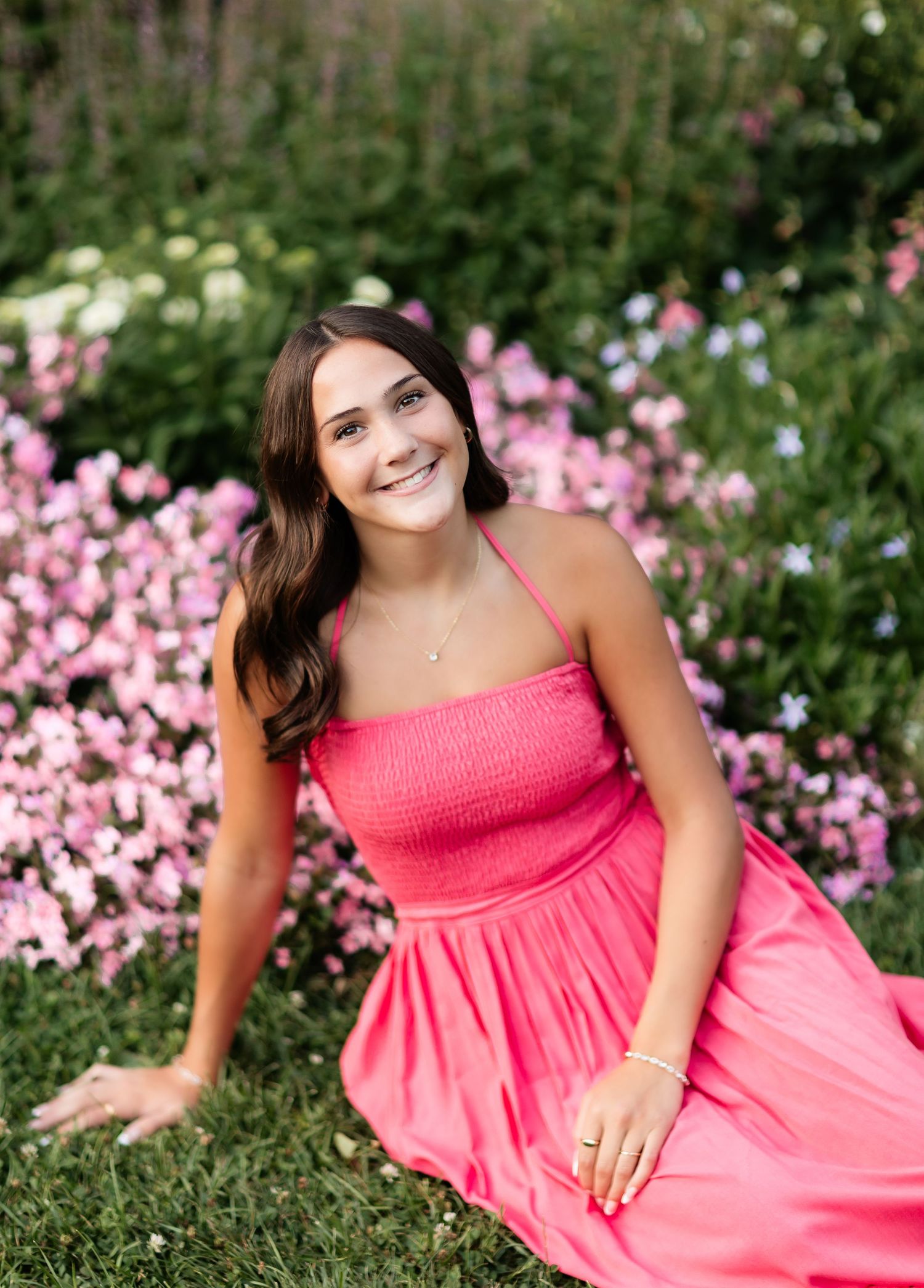 Young woman in pink halter dress sitting among colorful garden flowers, smiling outdoors.