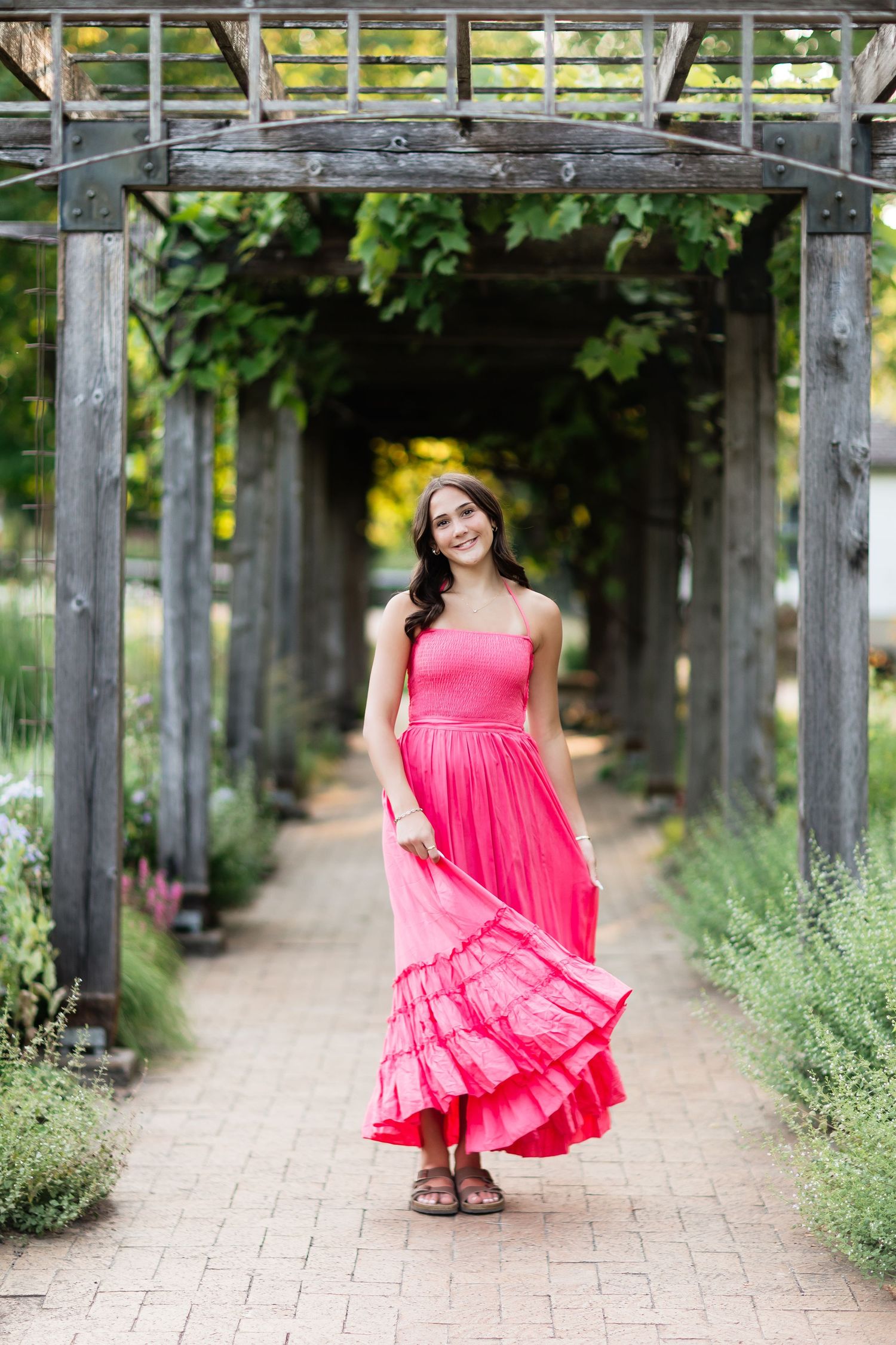 Young woman in pink maxi dress twirling under a wooden pergola surrounded by lush greenery in a garden.