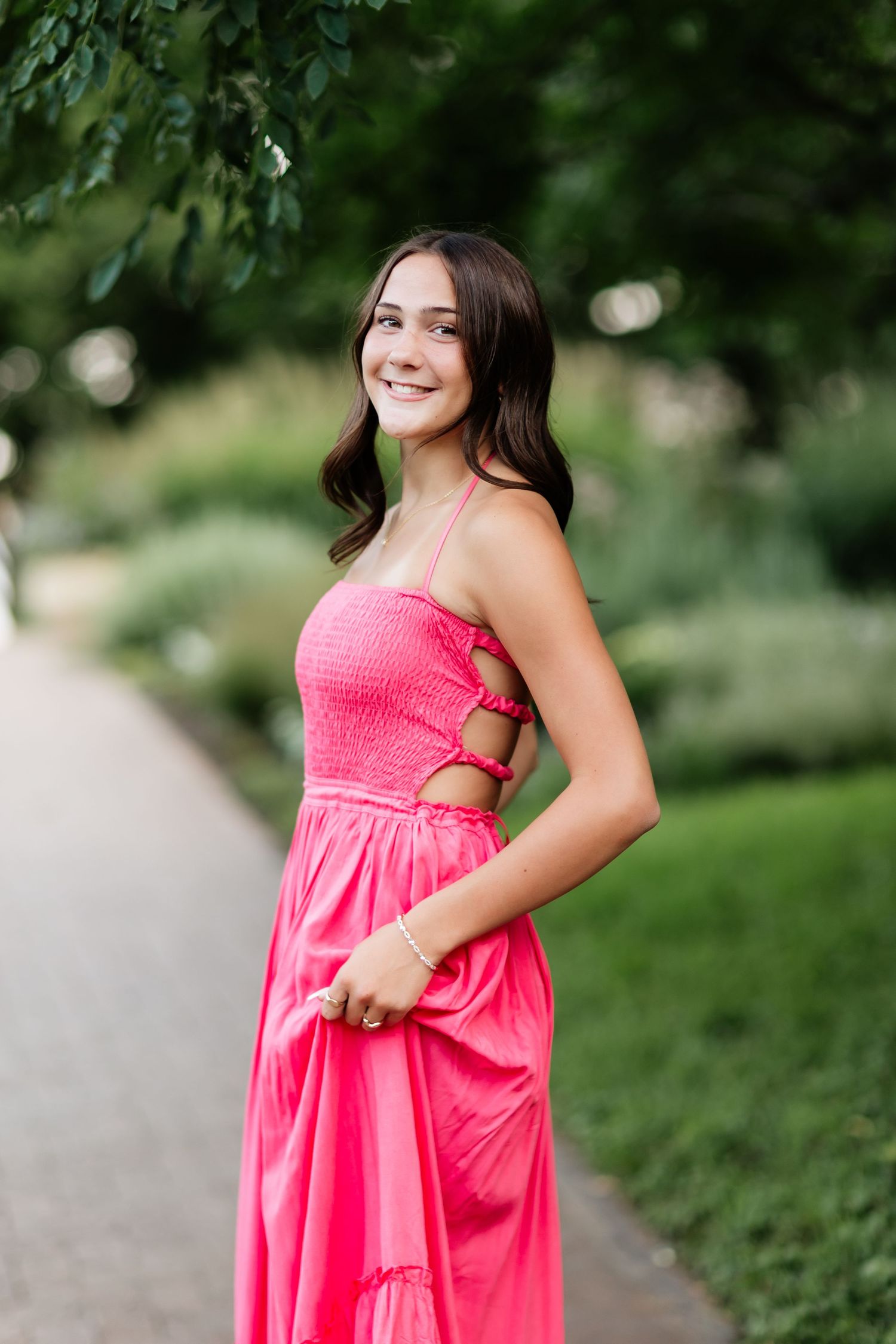 Smiling brunette teen in pink dress posing on a garden path among green trees.