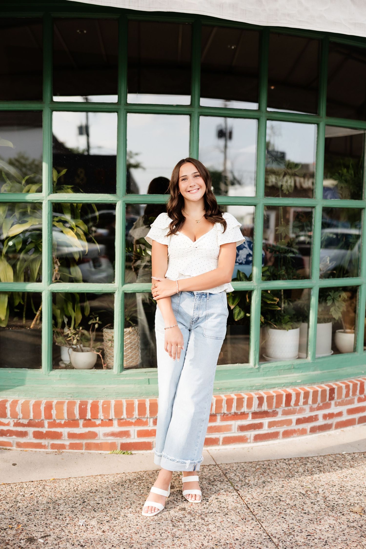 Young woman in white top and light jeans smiling outdoors by green storefront with plants.