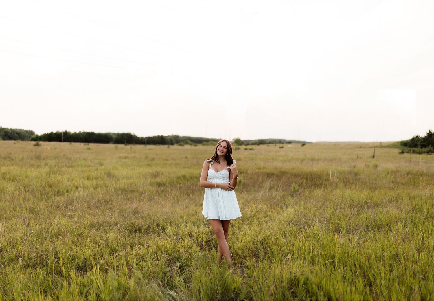 Young woman in white mini dress standing in a wide open grassy meadow under an overcast sky.