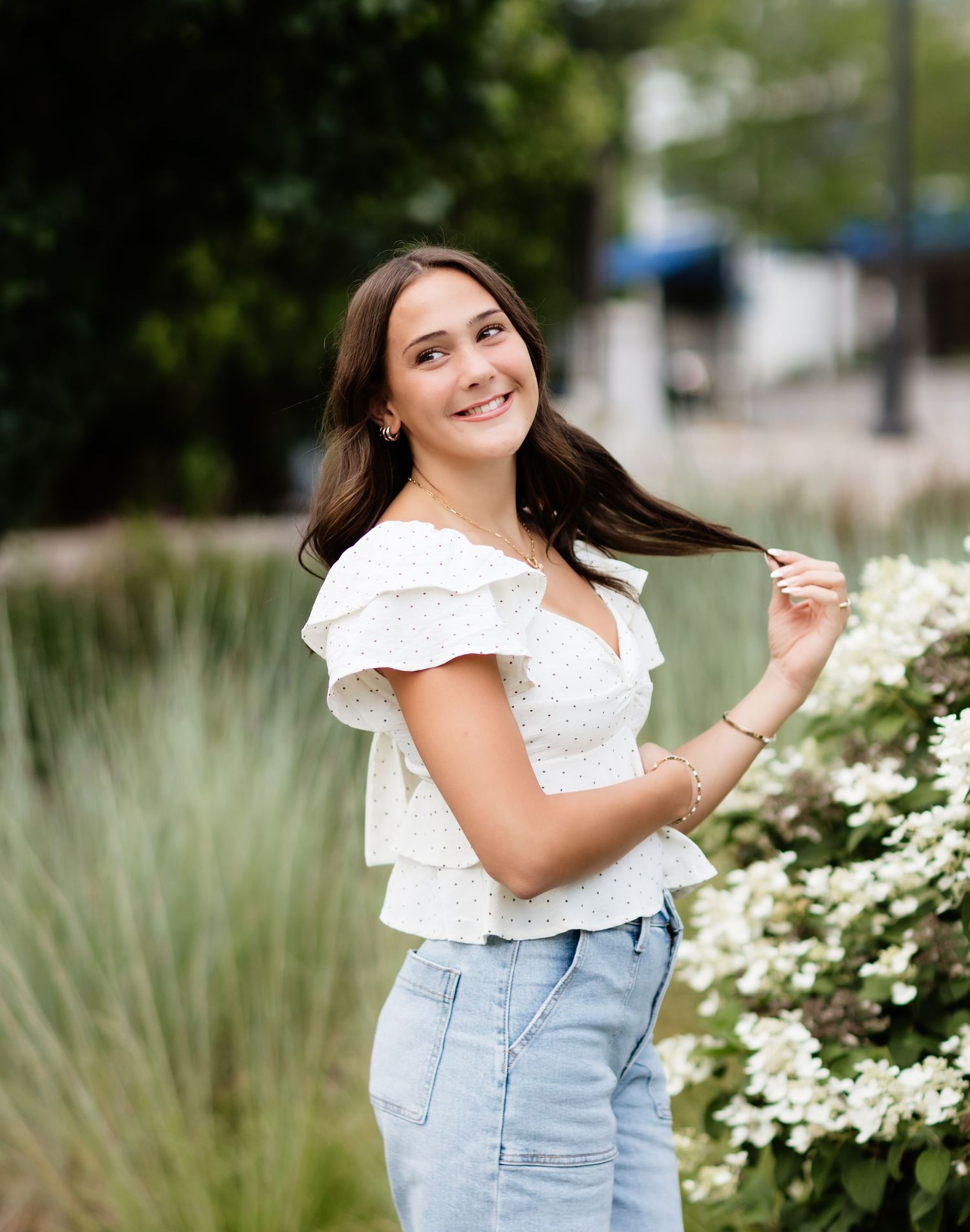 Young woman in white ruffled top and jeans smiling outdoors near white flowers and greenery.