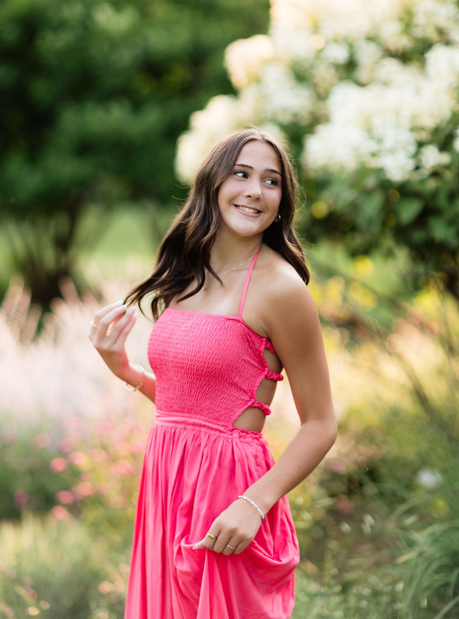 Young woman in bright pink smocked sundress poses among colorful garden flowers with a joyful smile.