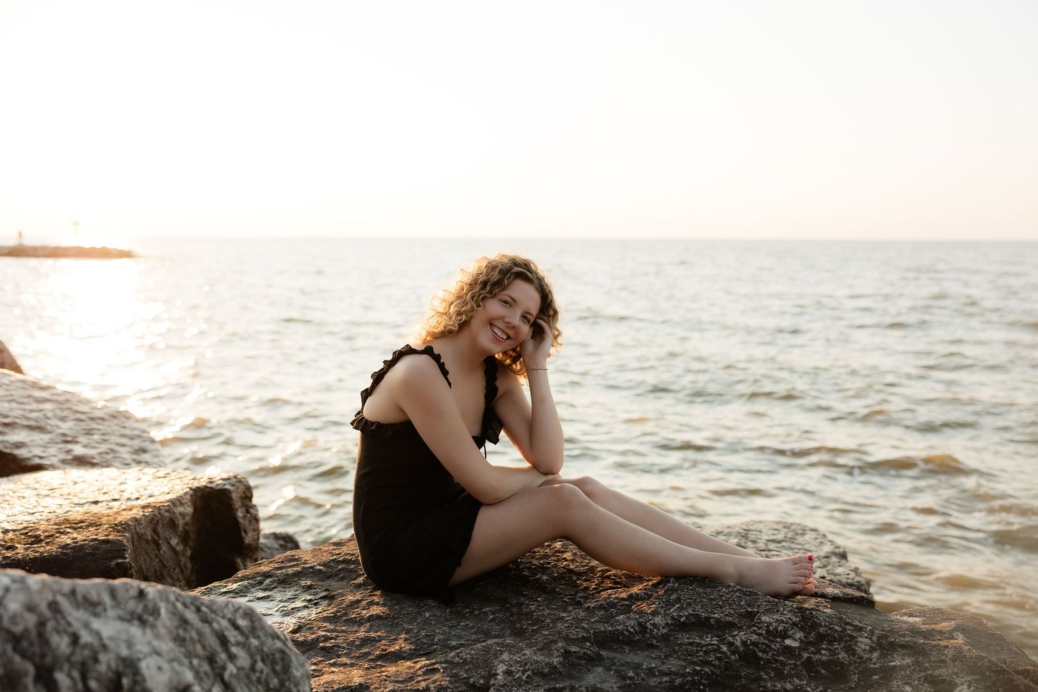 Young woman in black dress sitting on rocks by the sea at sunset, smiling peacefully.