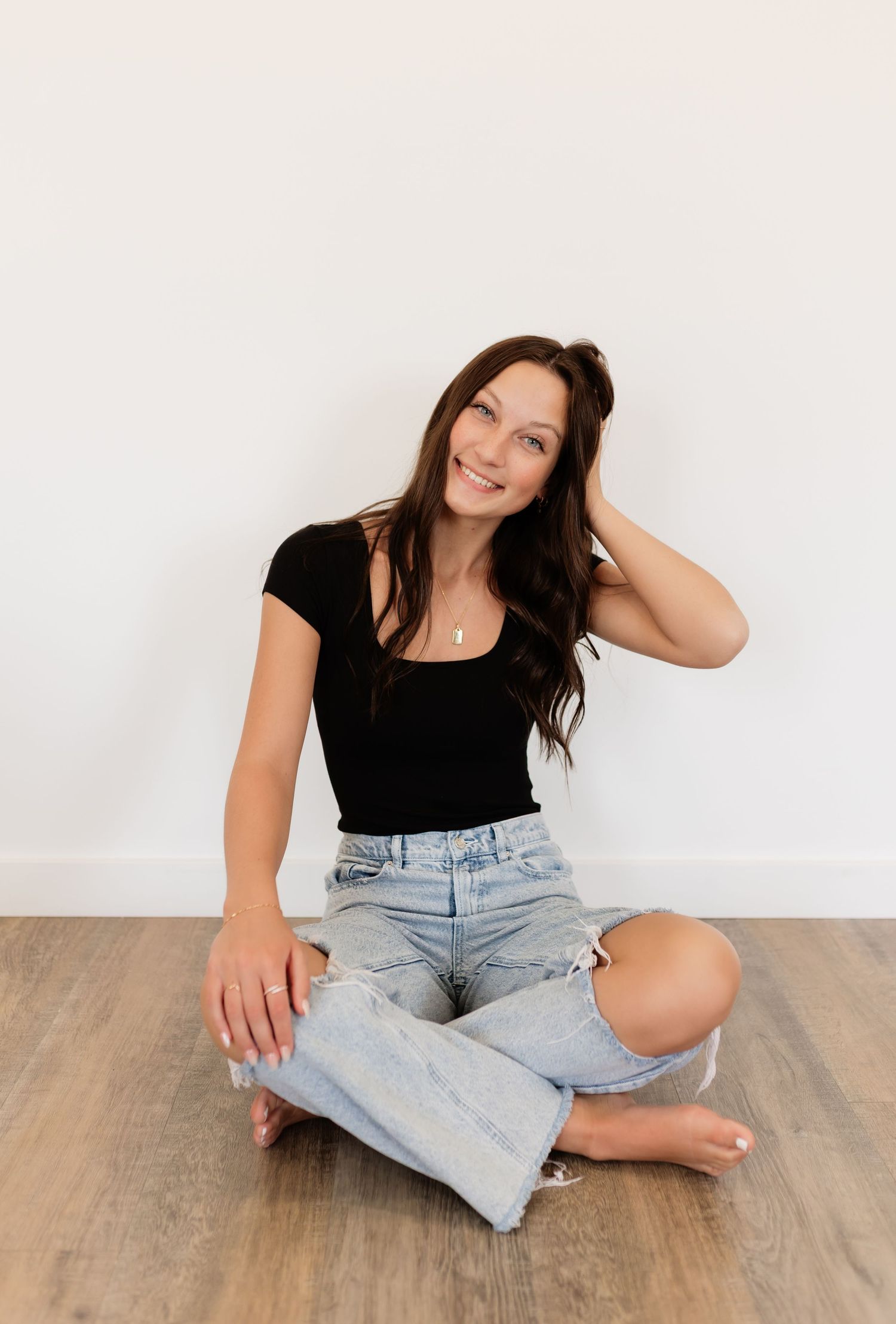 Brunette woman in black top and ripped jeans sitting cross-legged on wood floor.