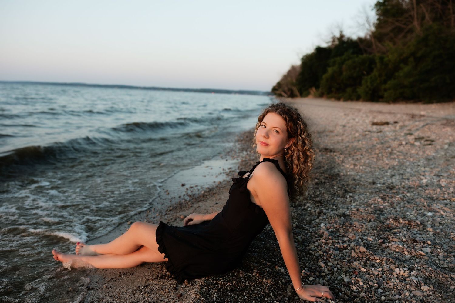 Young woman in black dress sitting on rocky beach shore at sunset with calm water and trees in background.