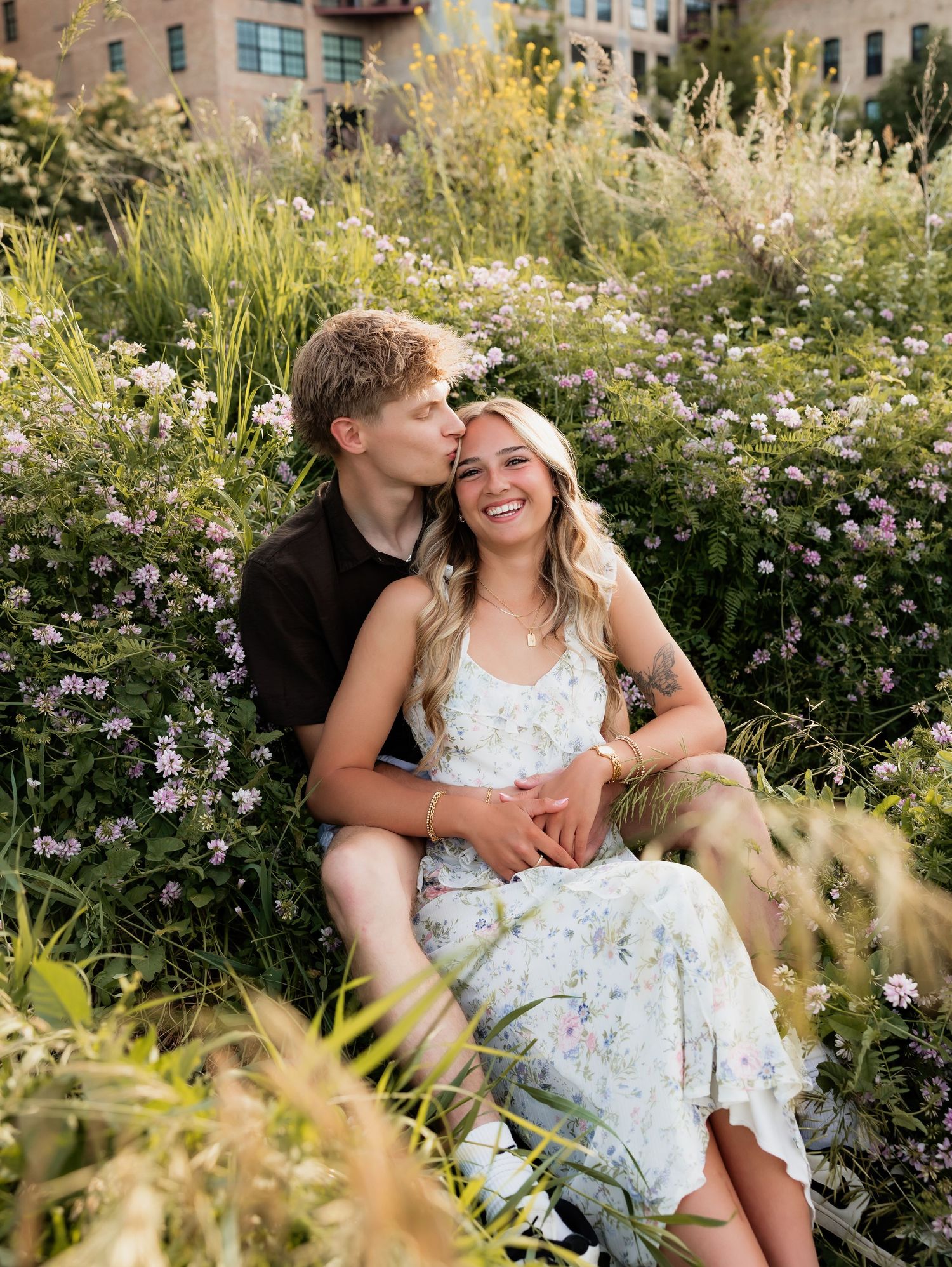 Happy couple sitting together in a wildflower meadow, man kissing woman's cheek as she smiles joyfully.