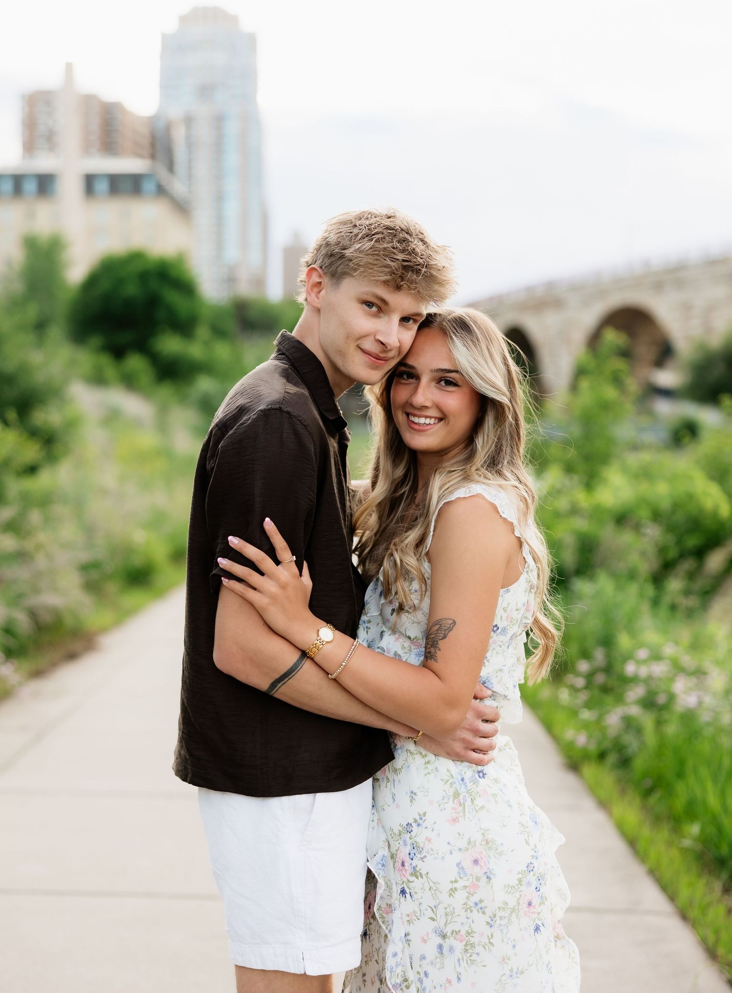 Happy young couple hugging on garden path, stone bridge and city skyline in background, smiling at camera.