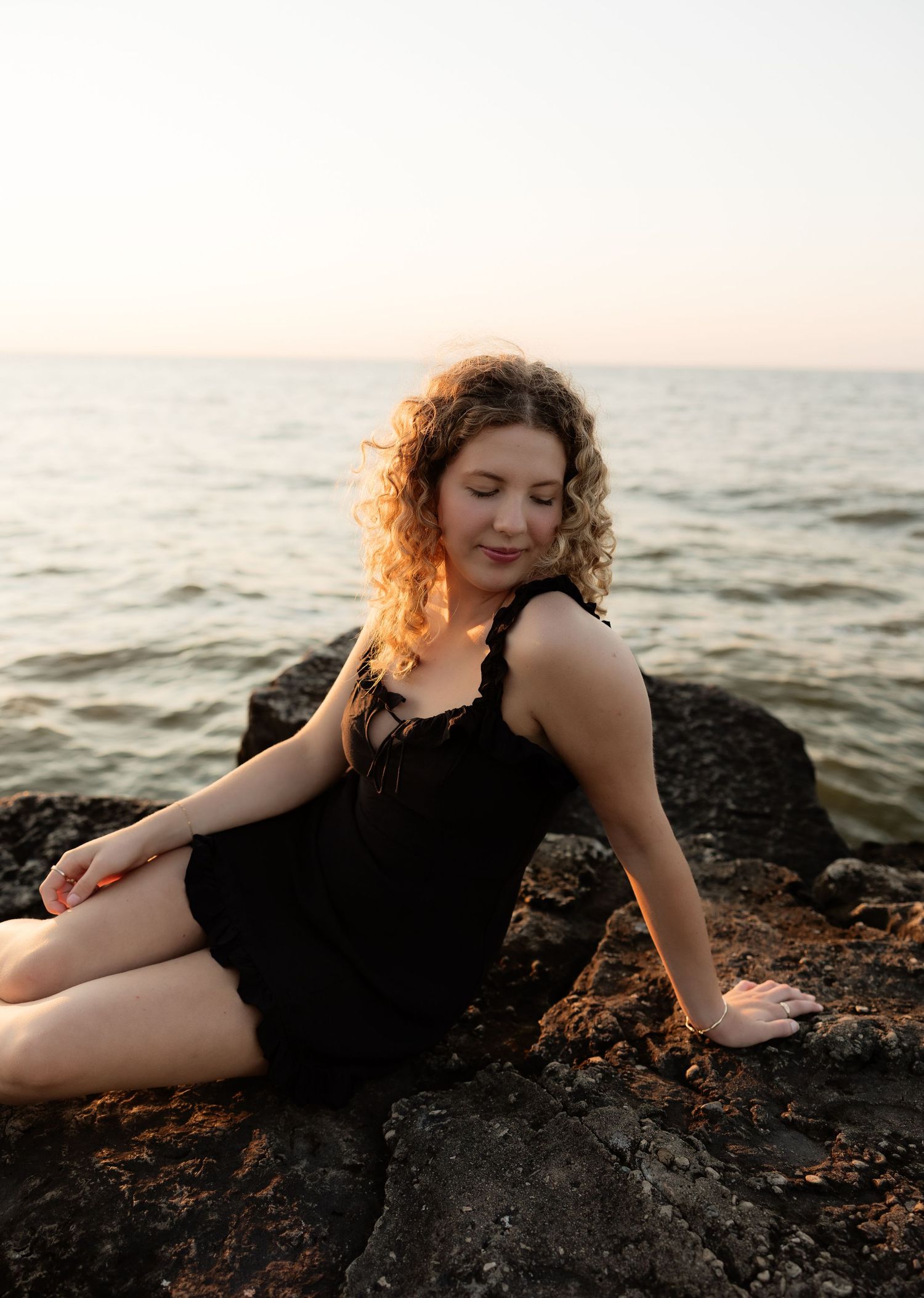 Young woman with curly hair in black dress sits on rocky shore at golden hour sunset by the ocean.