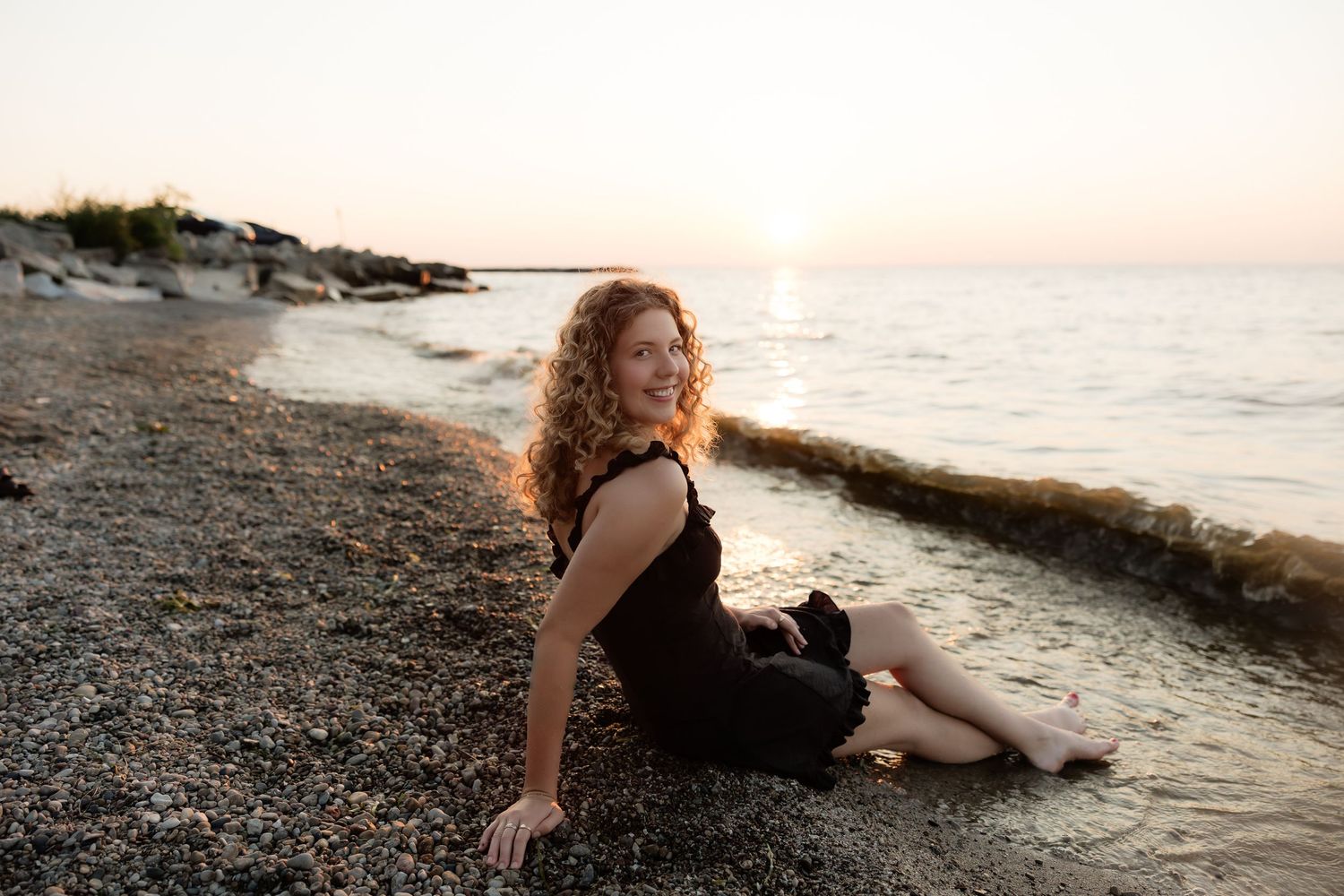 Smiling curly-haired woman in black dress sitting on pebble beach during golden hour sunset.