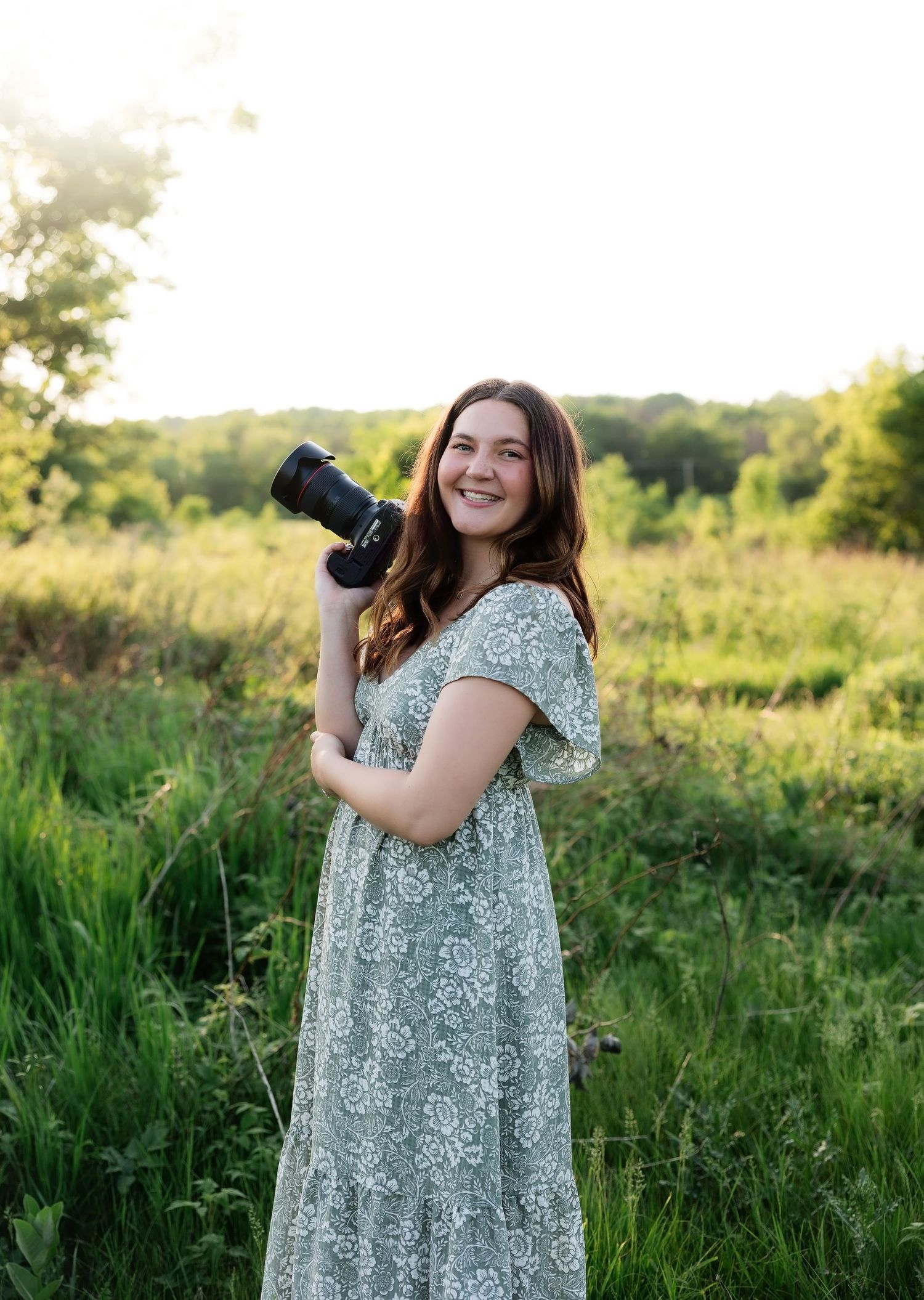 Young female photographer smiling in a floral maxi dress holding a camera in a sunlit green field.