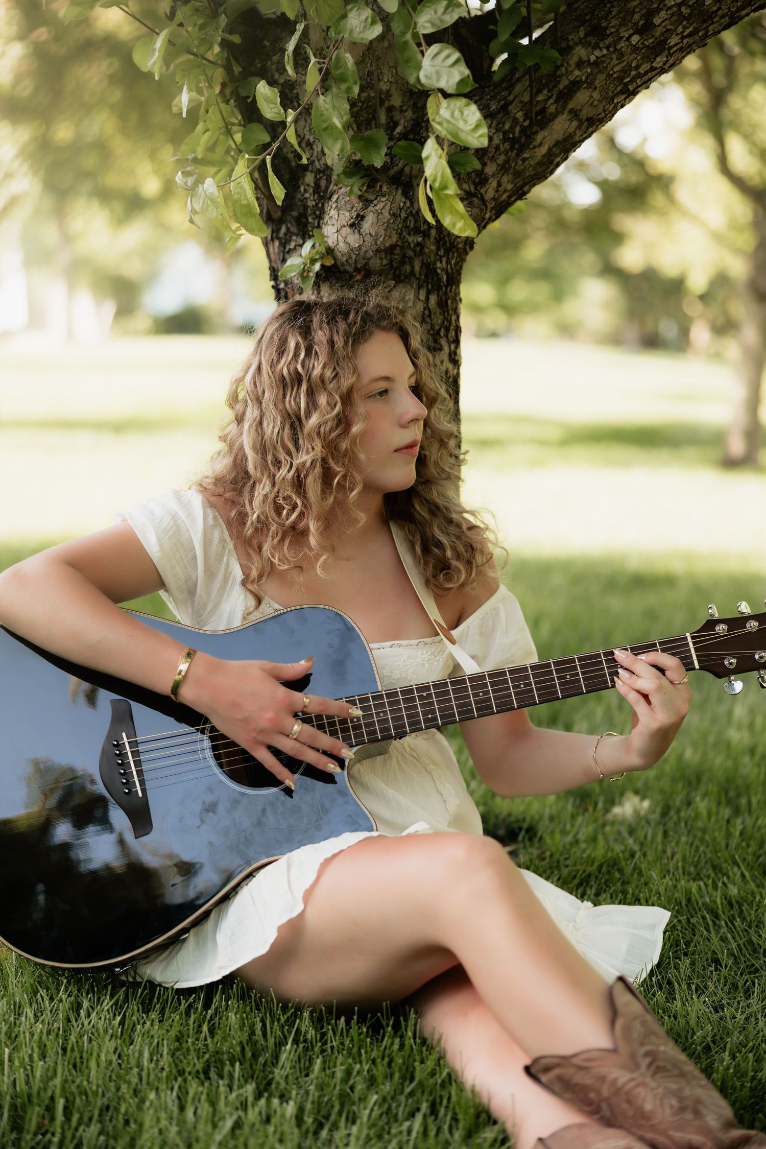 Young woman in white dress and cowboy boots playing acoustic guitar while sitting on grass under a tree.