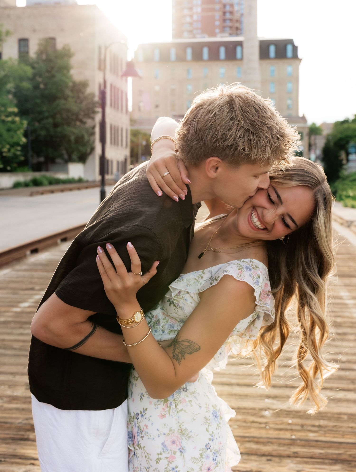 A joyful couple embracing outdoors, woman laughing as man kisses her cheek, wearing floral dress and black shirt.