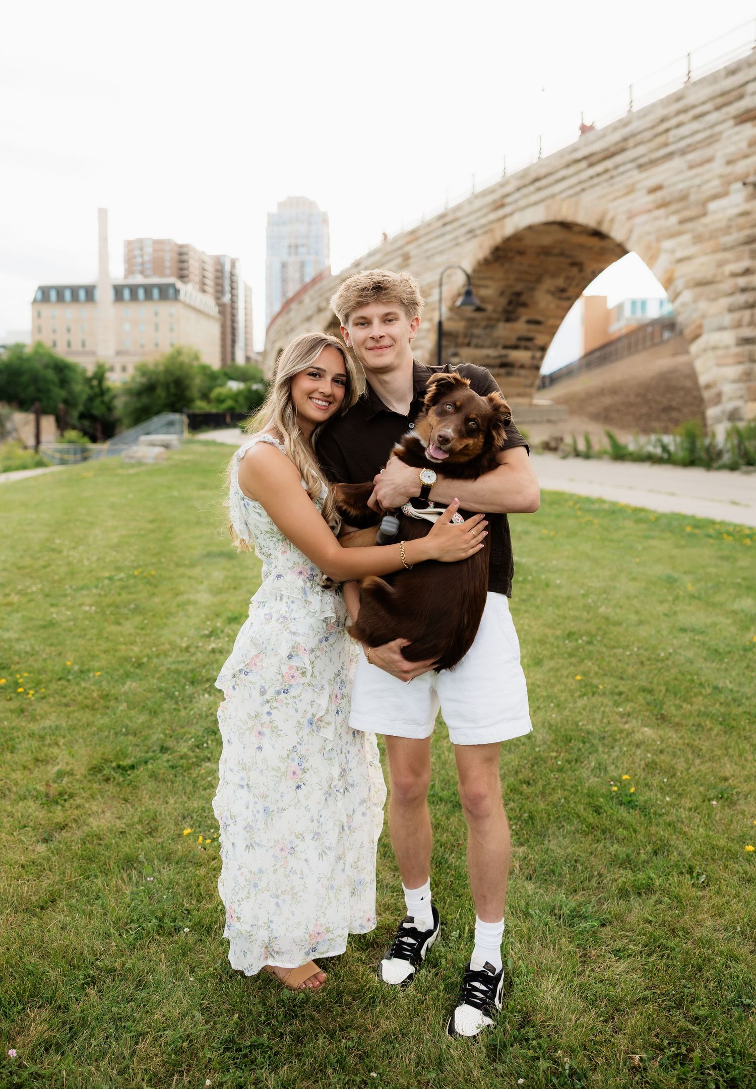Happy young couple holding a small brown dog near a stone arch bridge in a city park.