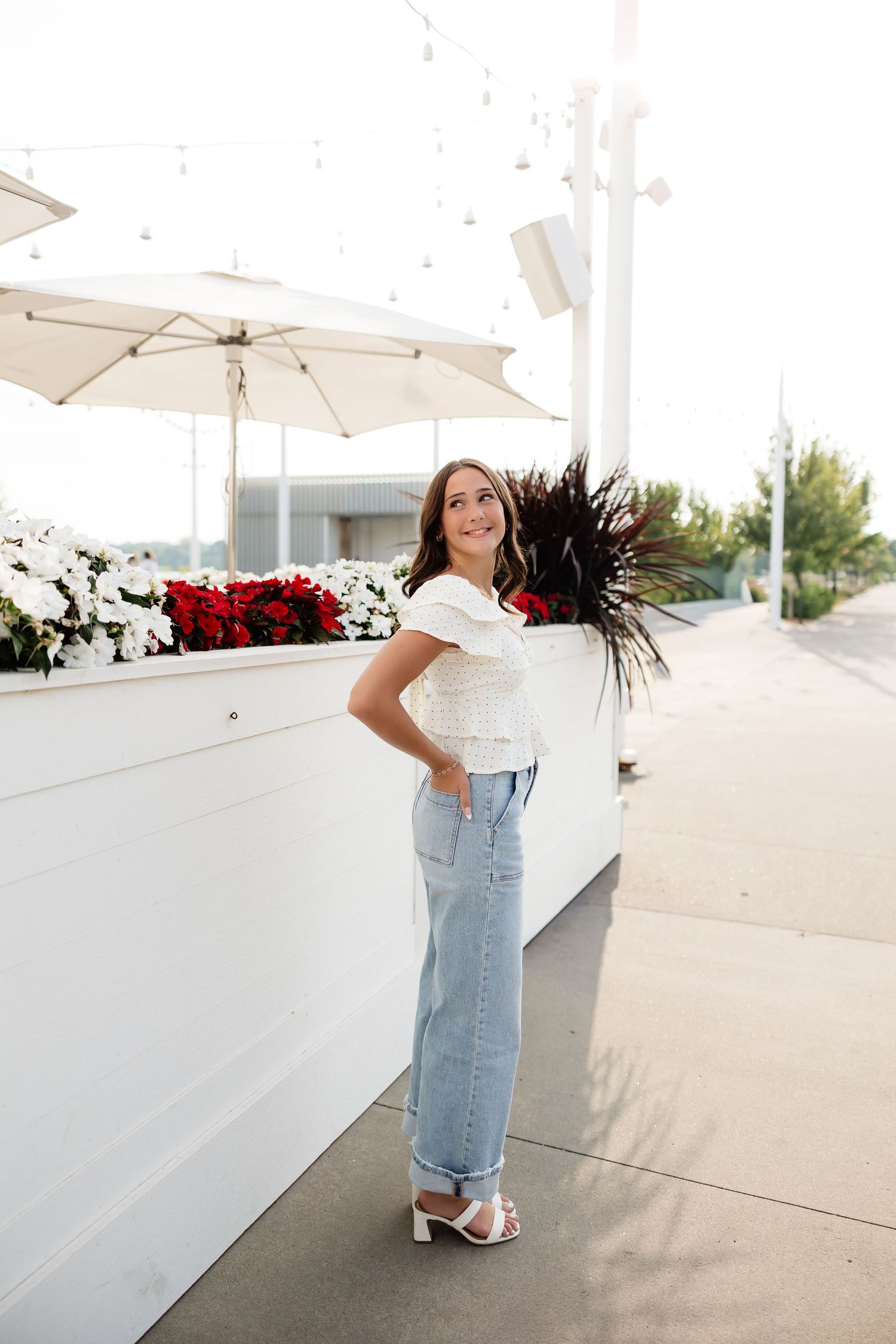 Young woman in white top and jeans posing outdoors near floral planter with red and white flowers.