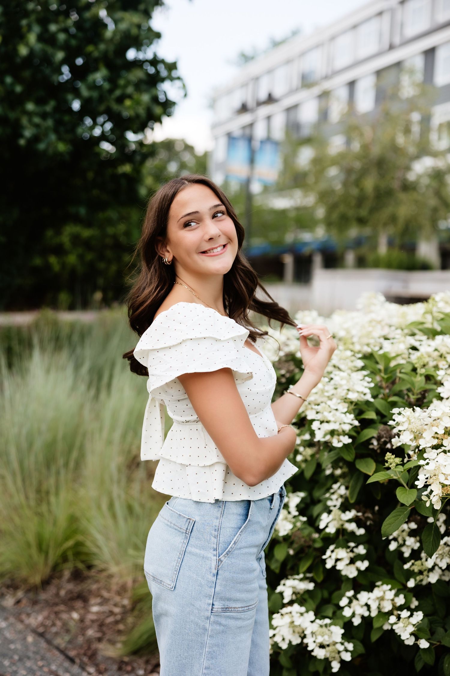 Smiling brunette woman in white eyelet top and light jeans posing by white hydrangea blooms.