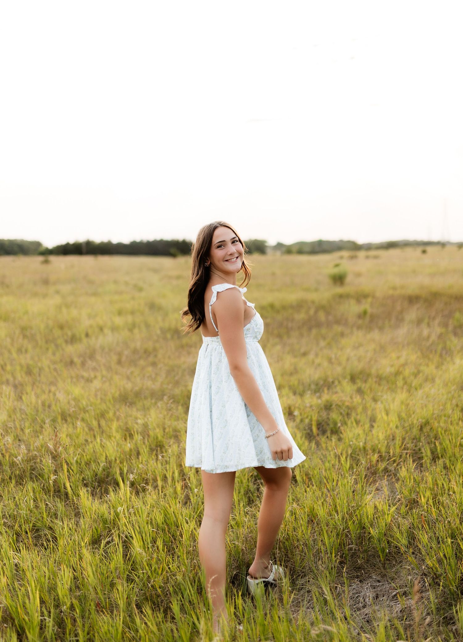 Young woman in white sundress smiling over shoulder in golden grass field.