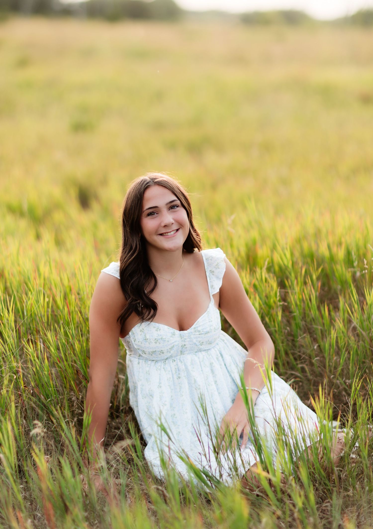 Smiling brunette girl in white dress sitting in a golden green grass field.