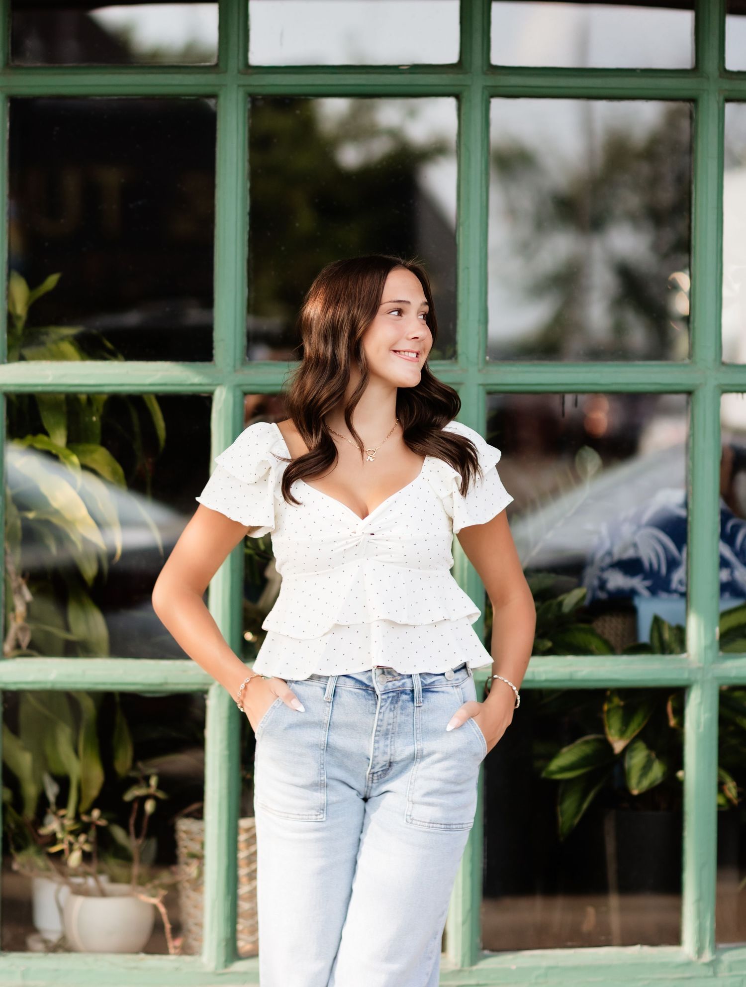Brunette woman in white tiered ruffle top and light jeans stands before green-framed window with plants.