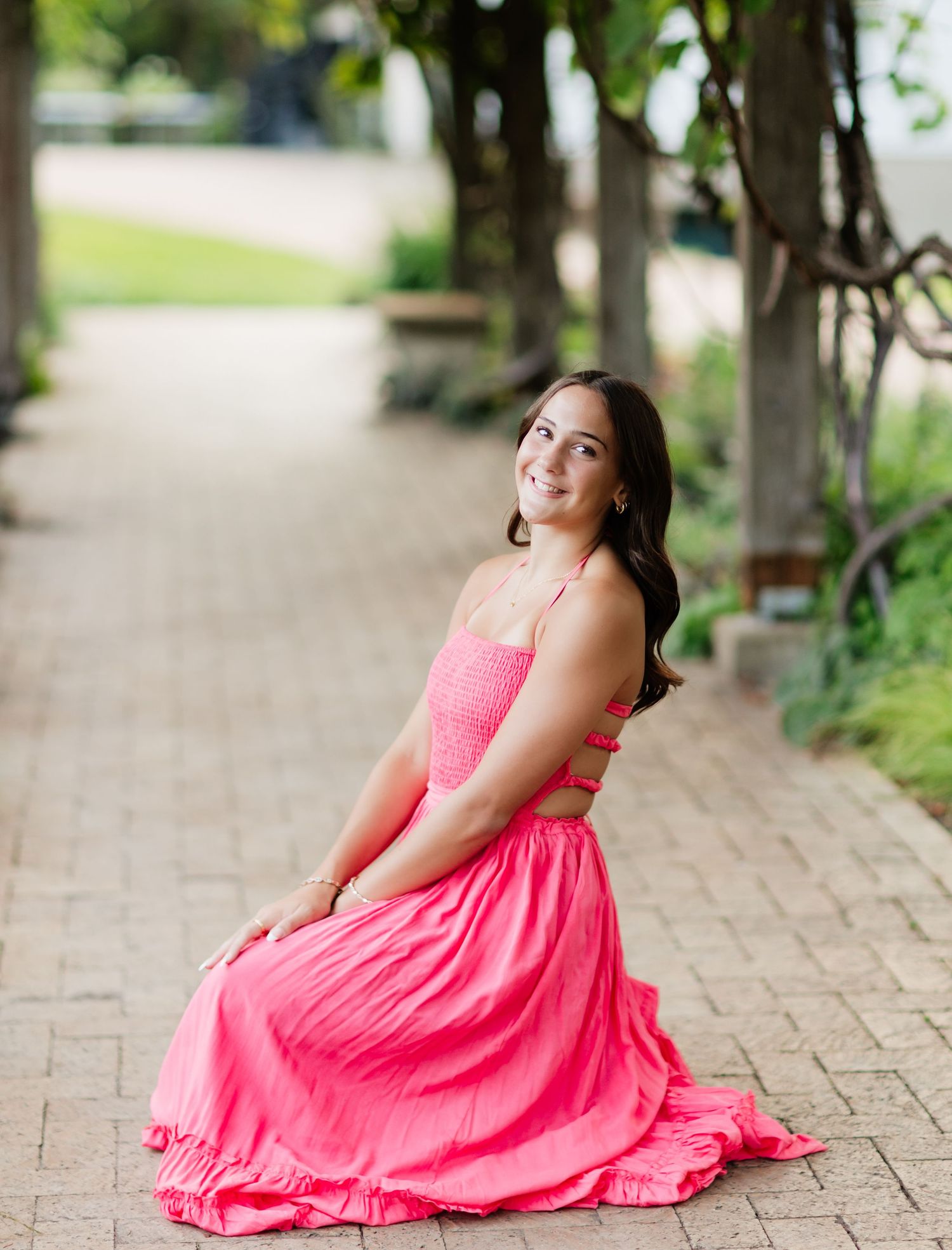Smiling young woman in pink maxi dress sitting on brick pathway surrounded by lush trees.