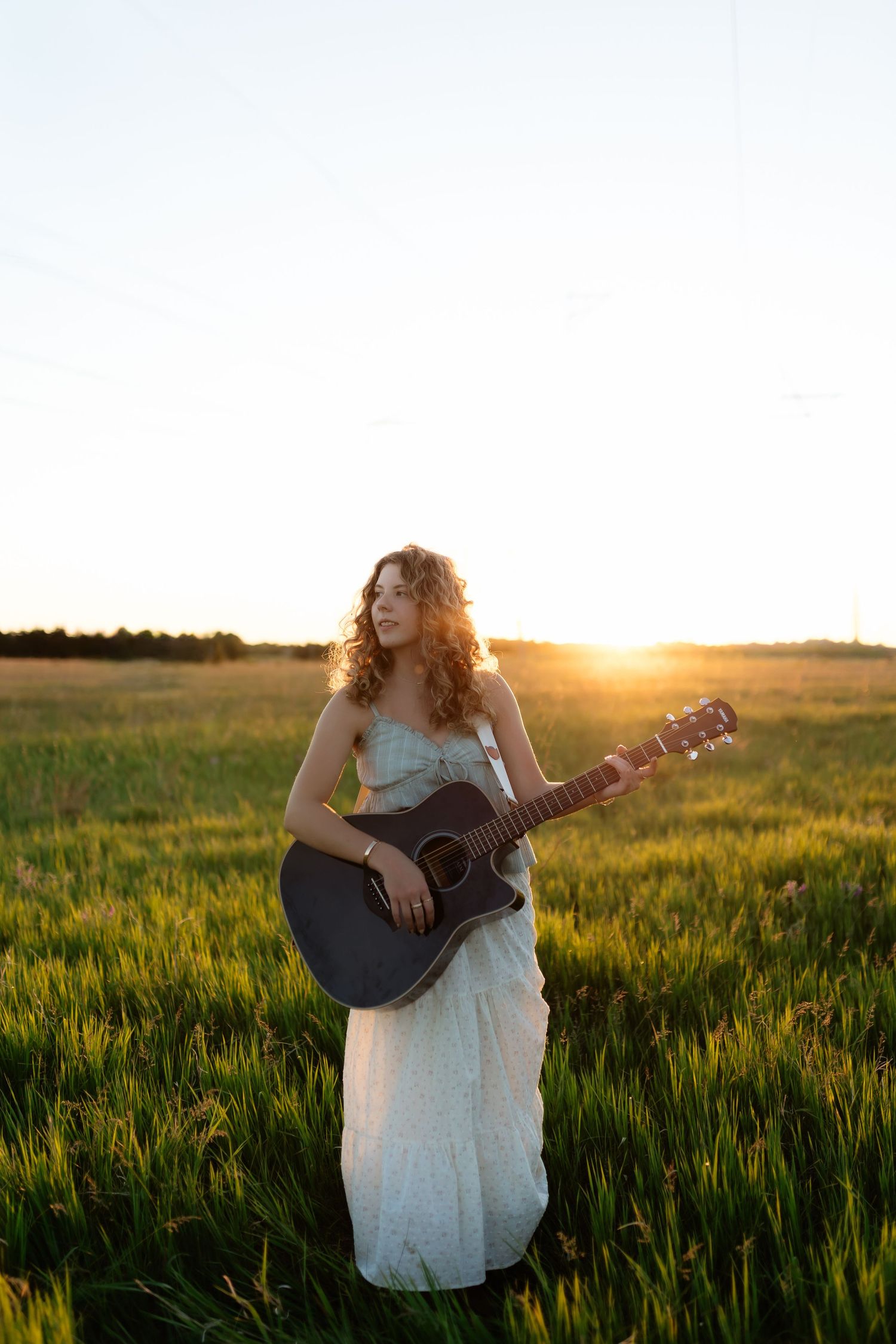 Young woman in light blue dress playing acoustic guitar while standing in a green field at sunset.
