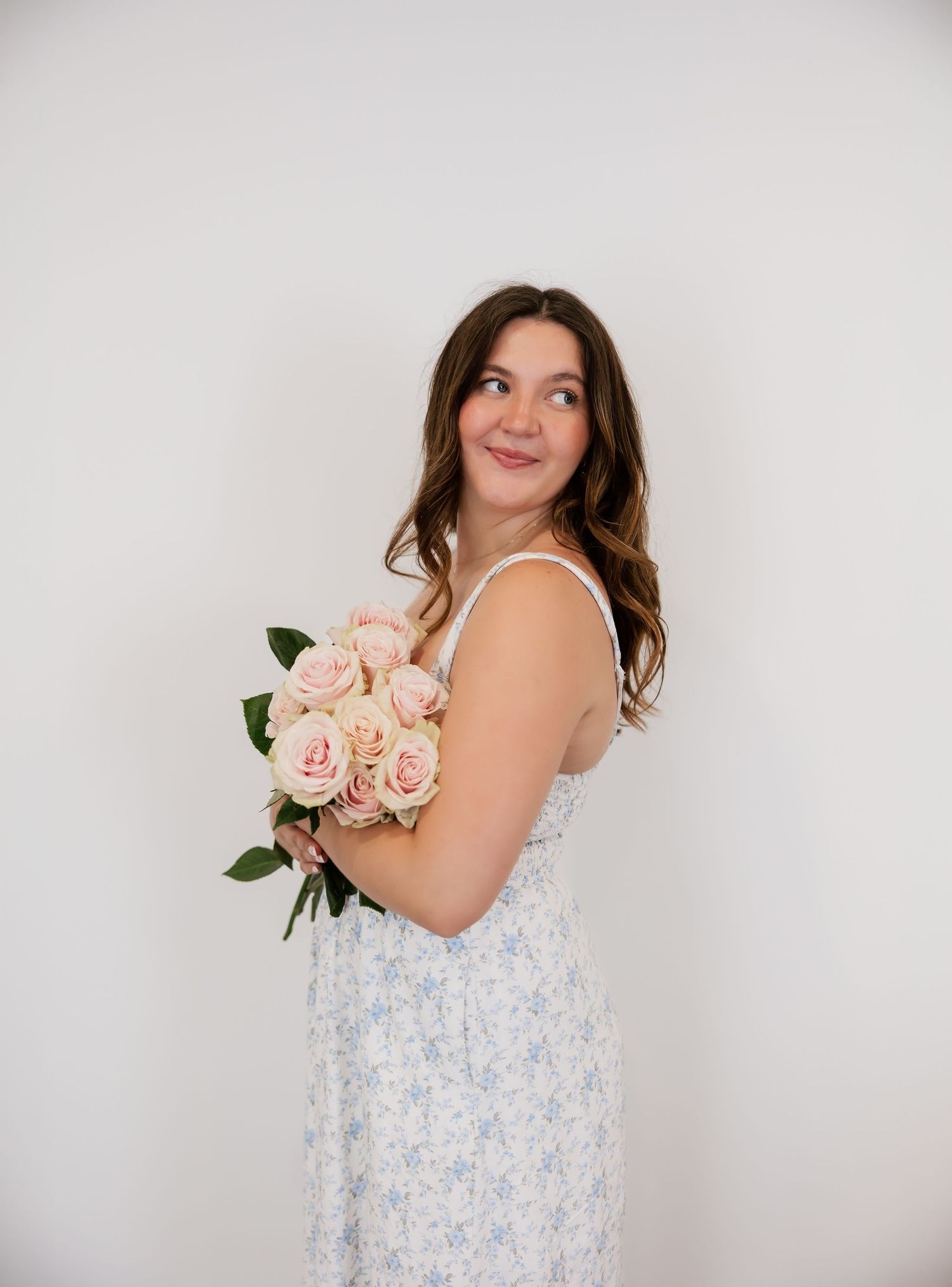 Young woman in floral blue dress holding pink roses bouquet, smiling over shoulder against white background.