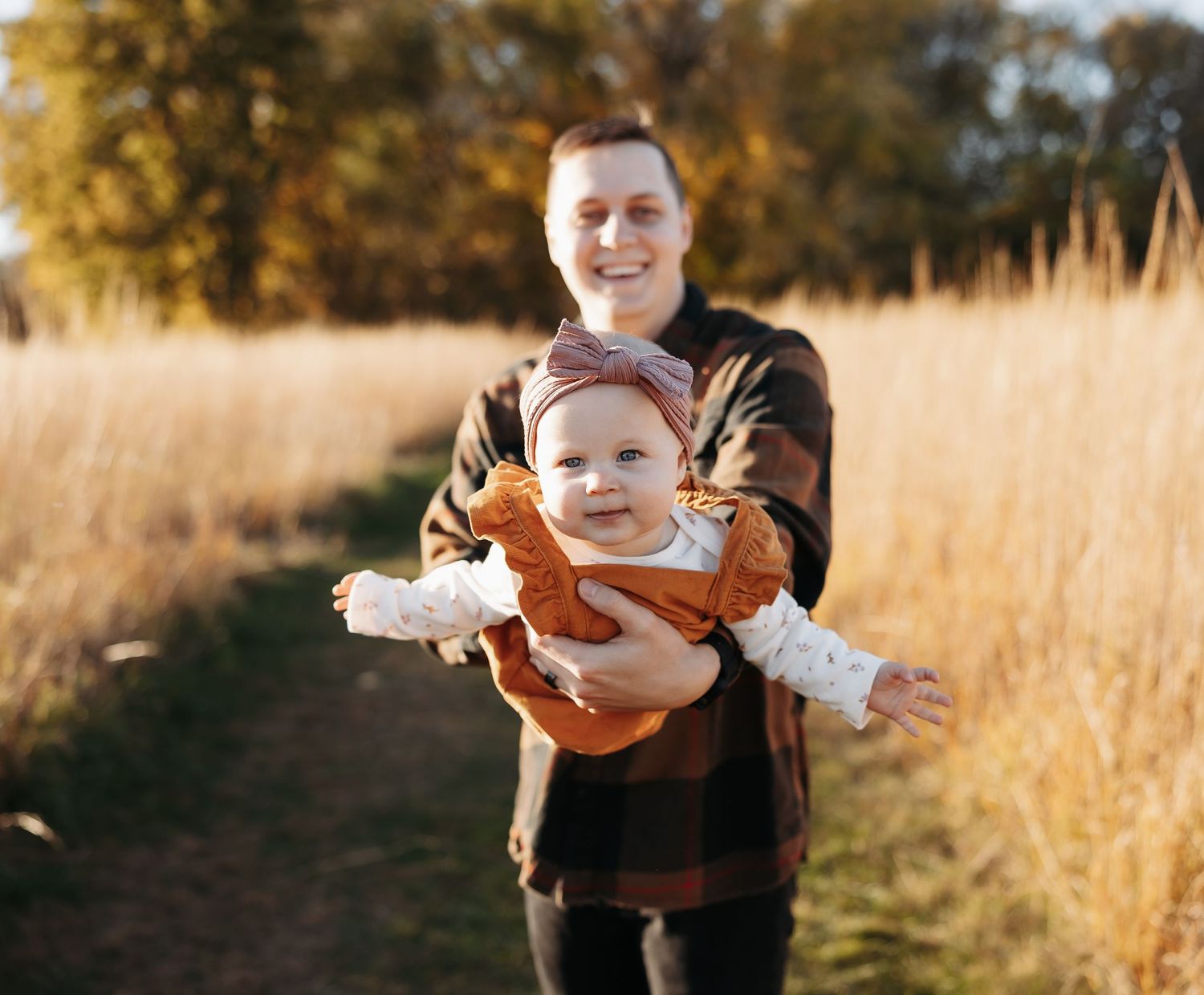 Smiling father holding a laughing baby girl in rust orange outfit in a golden autumn meadow.