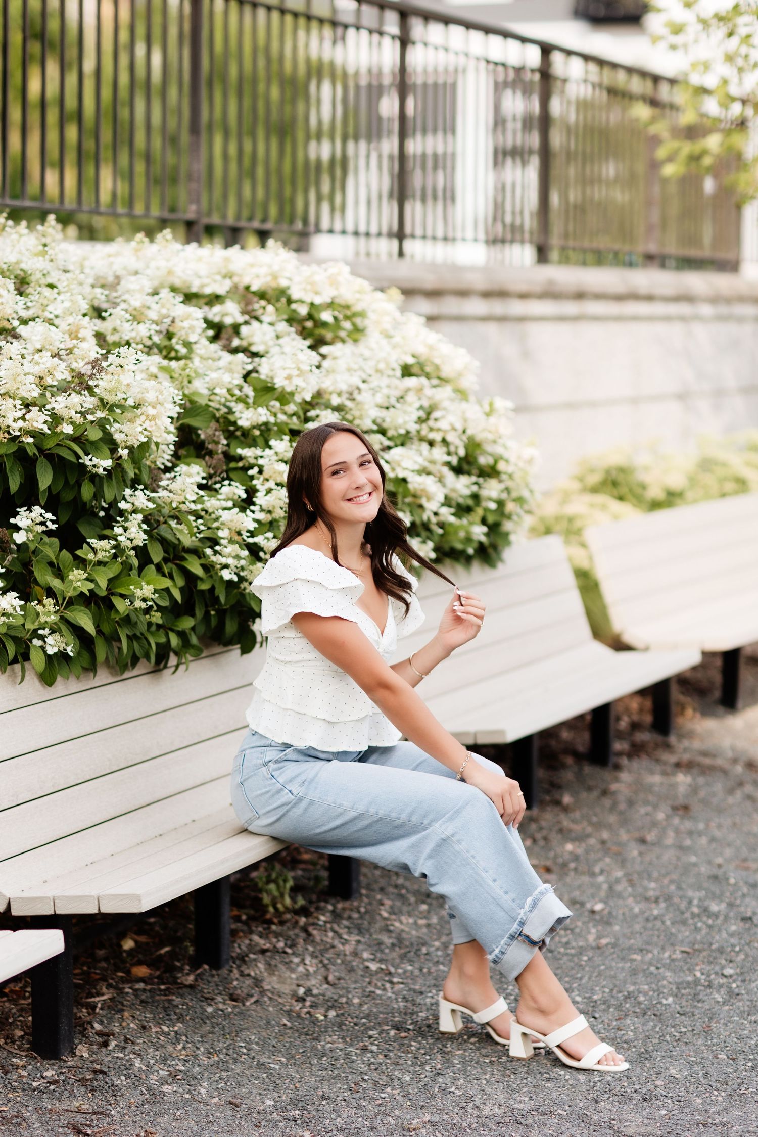 Smiling young woman in white ruffle top and jeans sitting on wooden bench beside white hydrangea bushes.