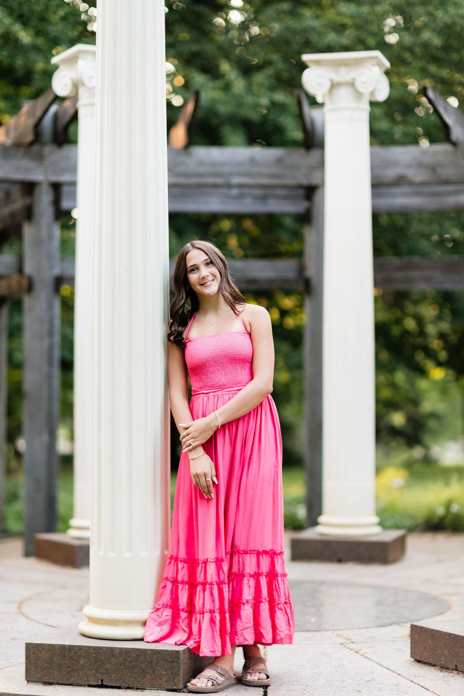 Smiling young woman in a pink maxi dress stands gracefully beside white columns in a lush garden setting.