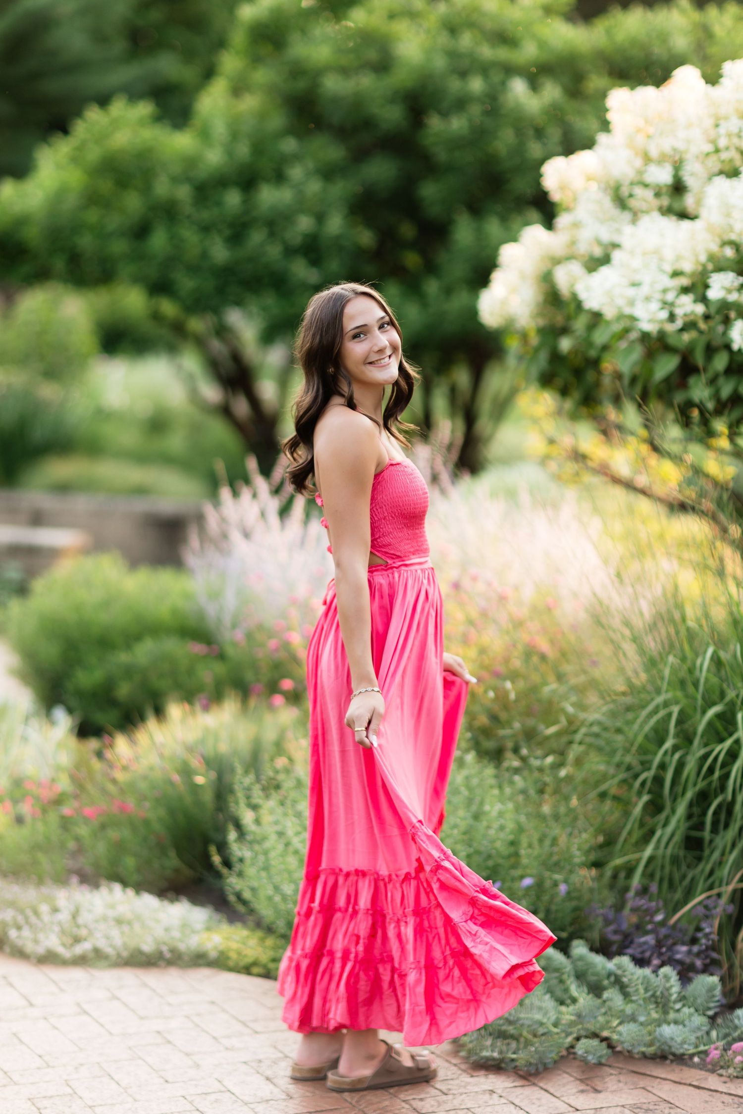 Young woman in a flowing pink maxi dress twirling on a brick path in a lush garden.
