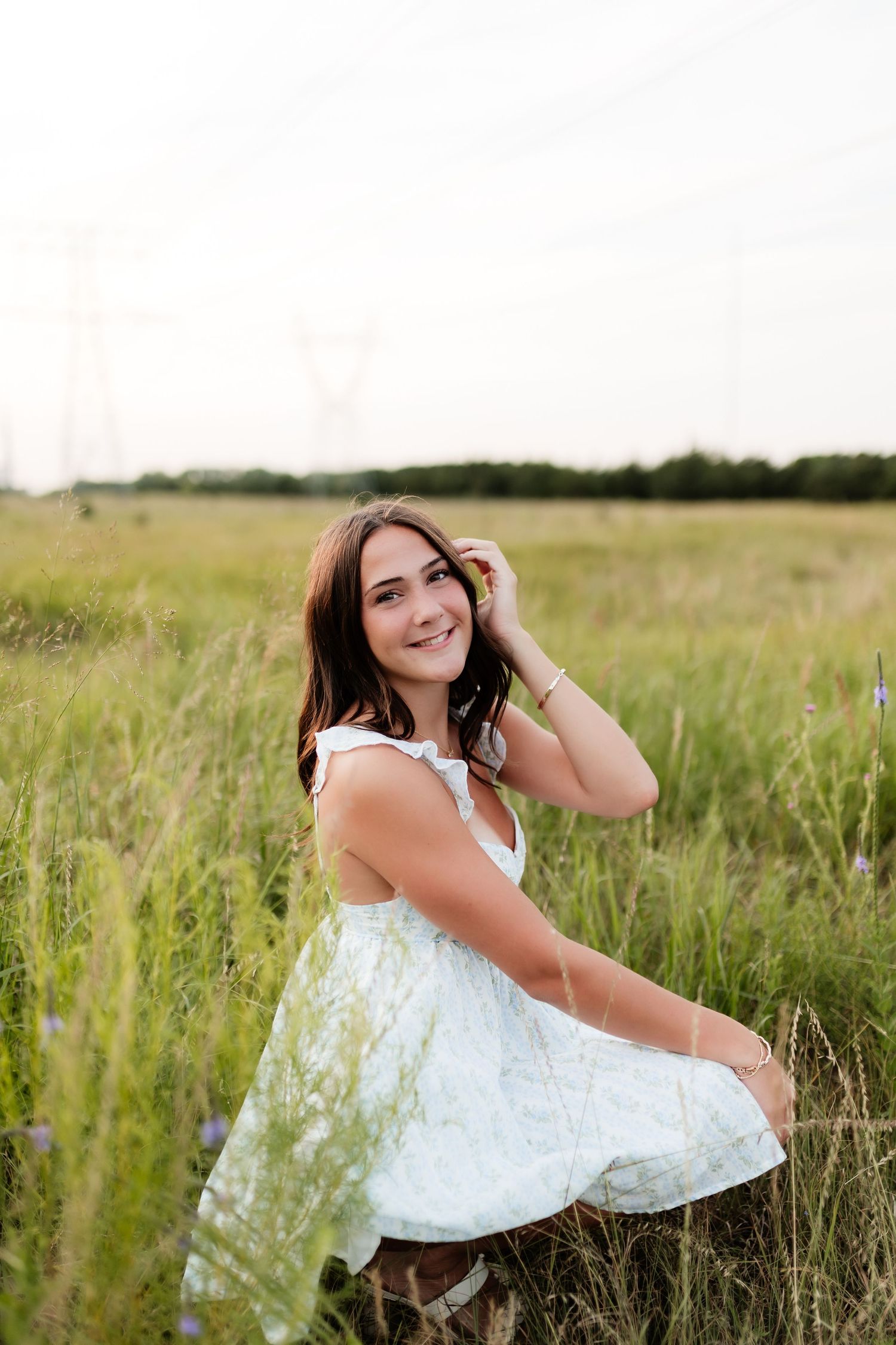Smiling young woman in white dress sitting in green meadow on bright day.