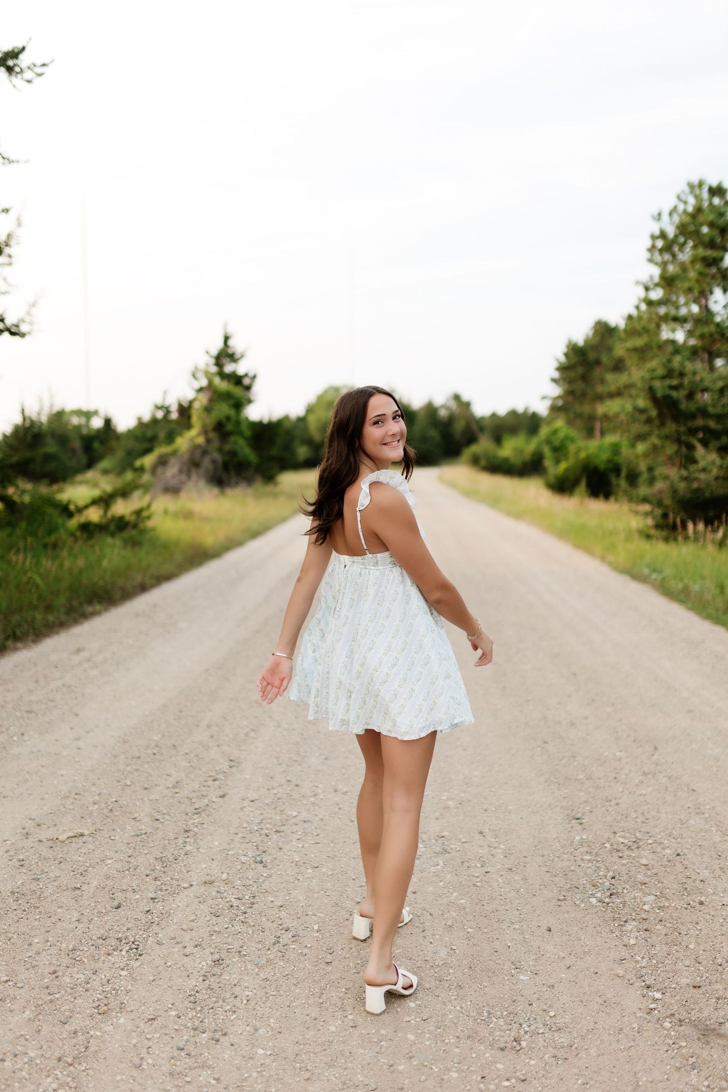 Smiling young woman in white sundress twirling on a peaceful gravel road surrounded by greenery.