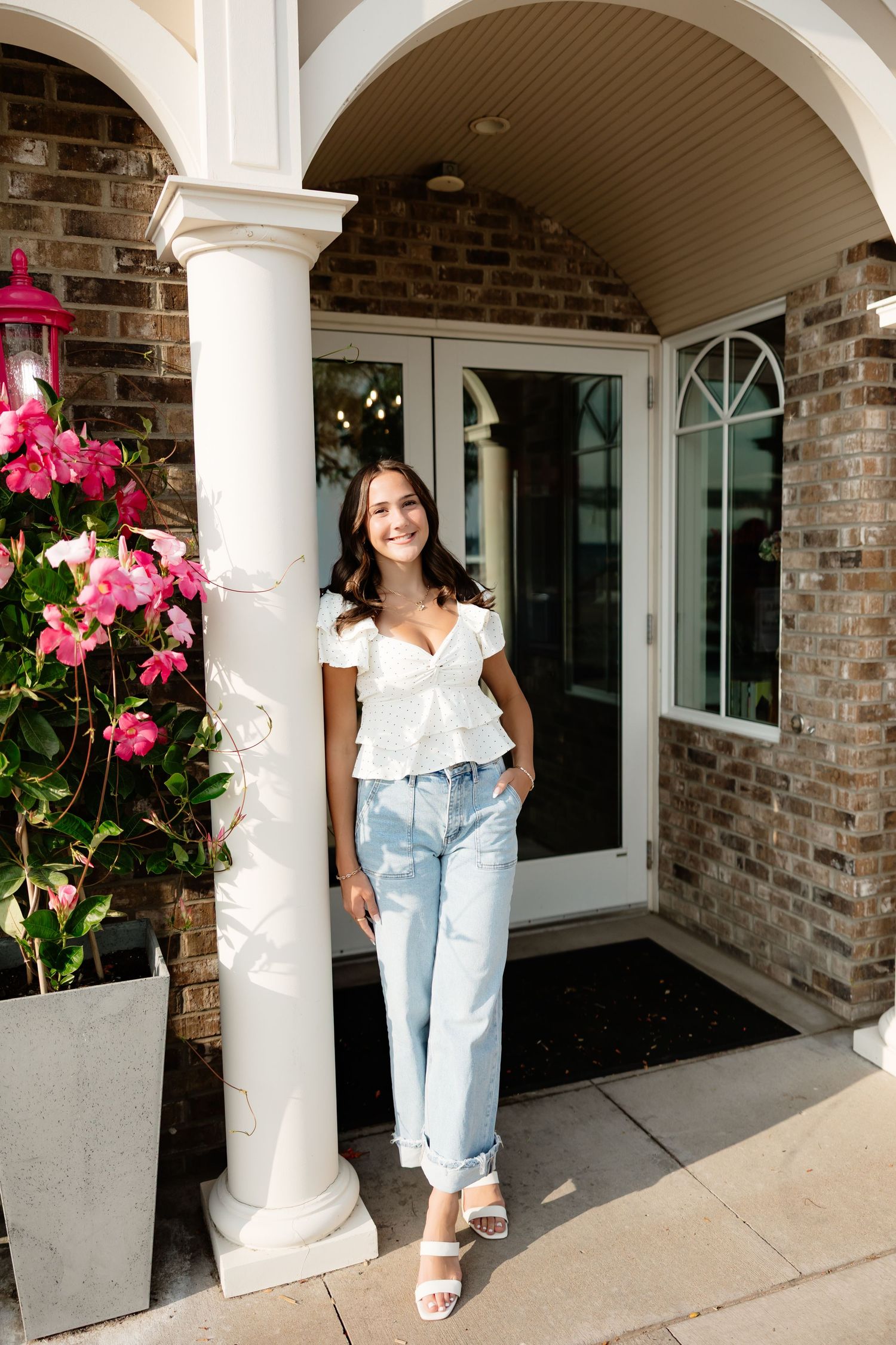 Smiling young woman in white top and jeans posing by column at brick building entrance.