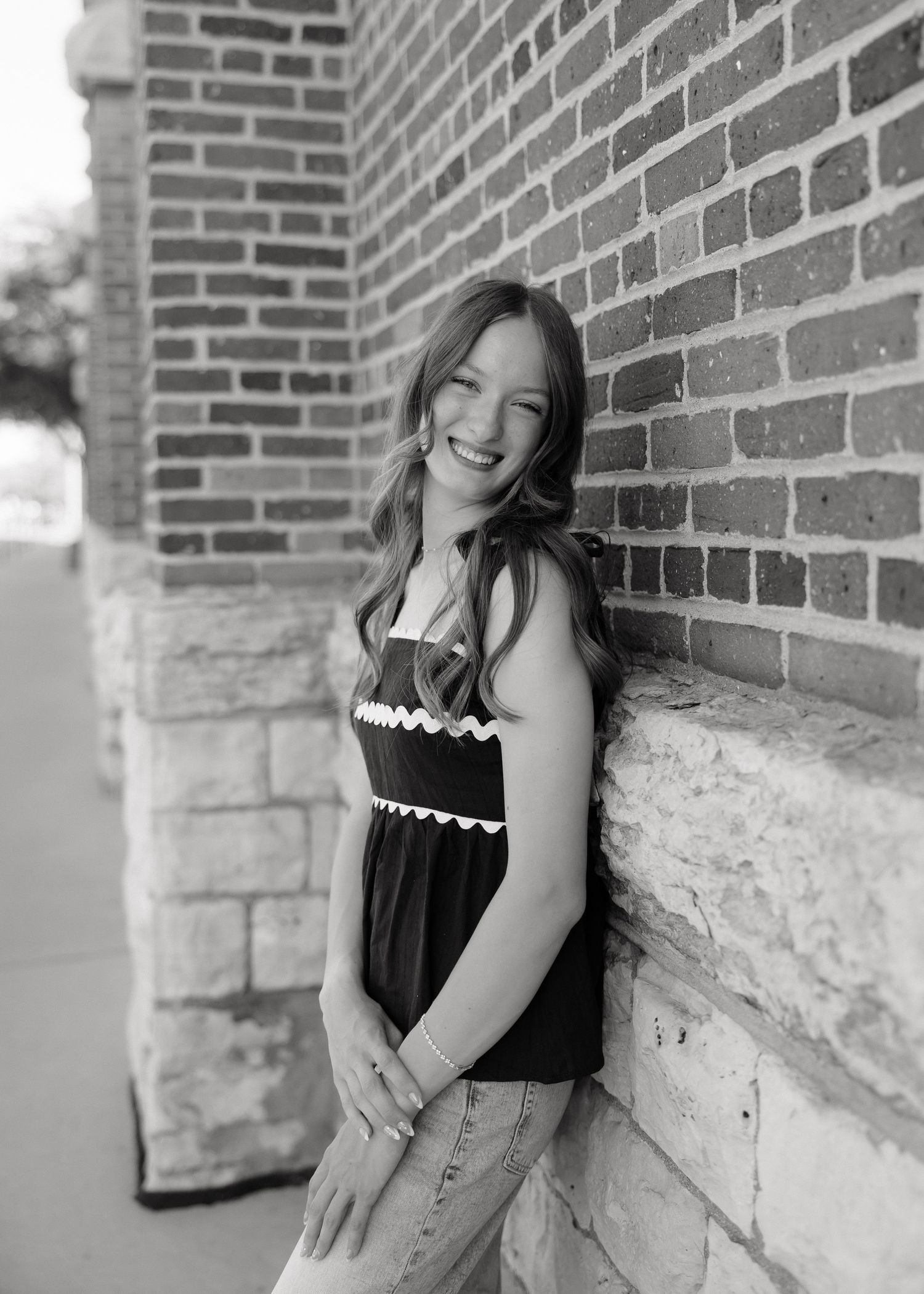Young woman smiling and leaning against a brick wall outdoors in a black and white portrait photo.