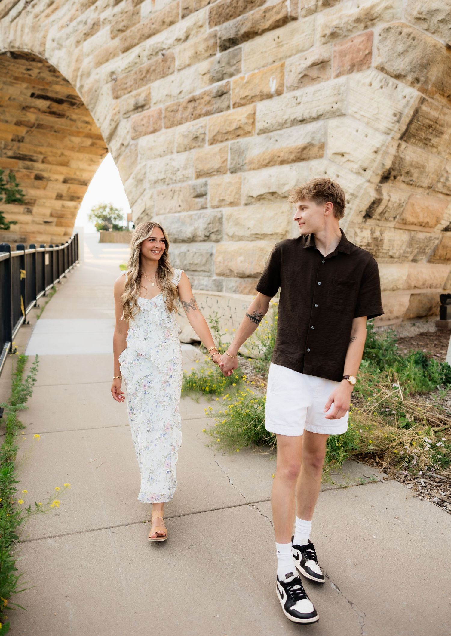 Couple holding hands walking under a stone arch bridge, woman in floral maxi dress, man in black shirt and white shorts.
