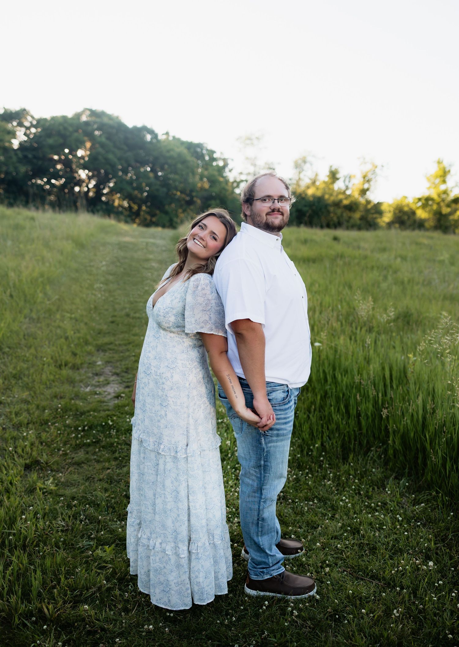 Couple posing back-to-back in a green meadow, woman in floral dress, man in white shirt and jeans, golden hour lighting.