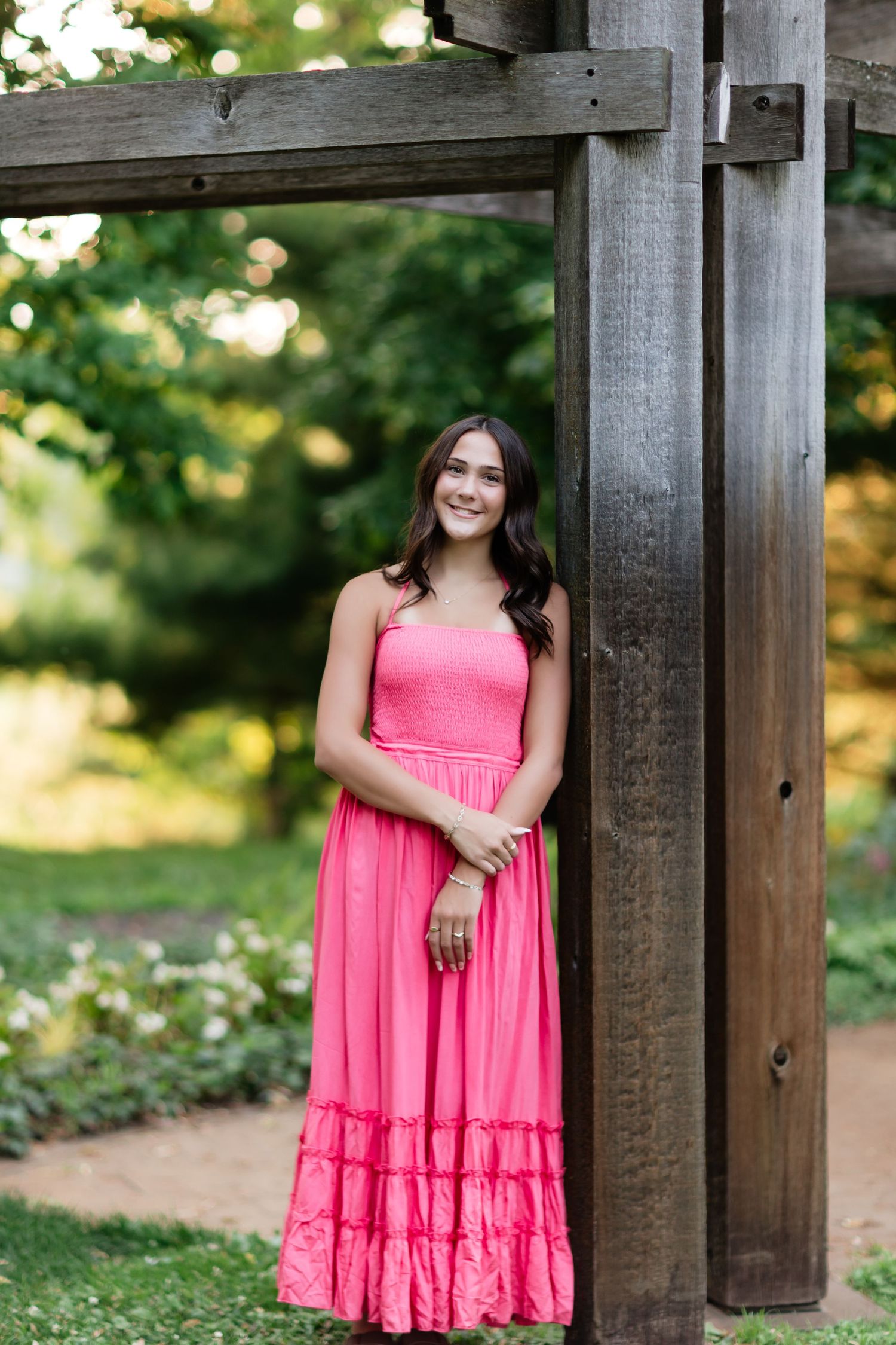 Young woman in a pink strapless maxi dress smiling while leaning against a rustic wooden structure outdoors.