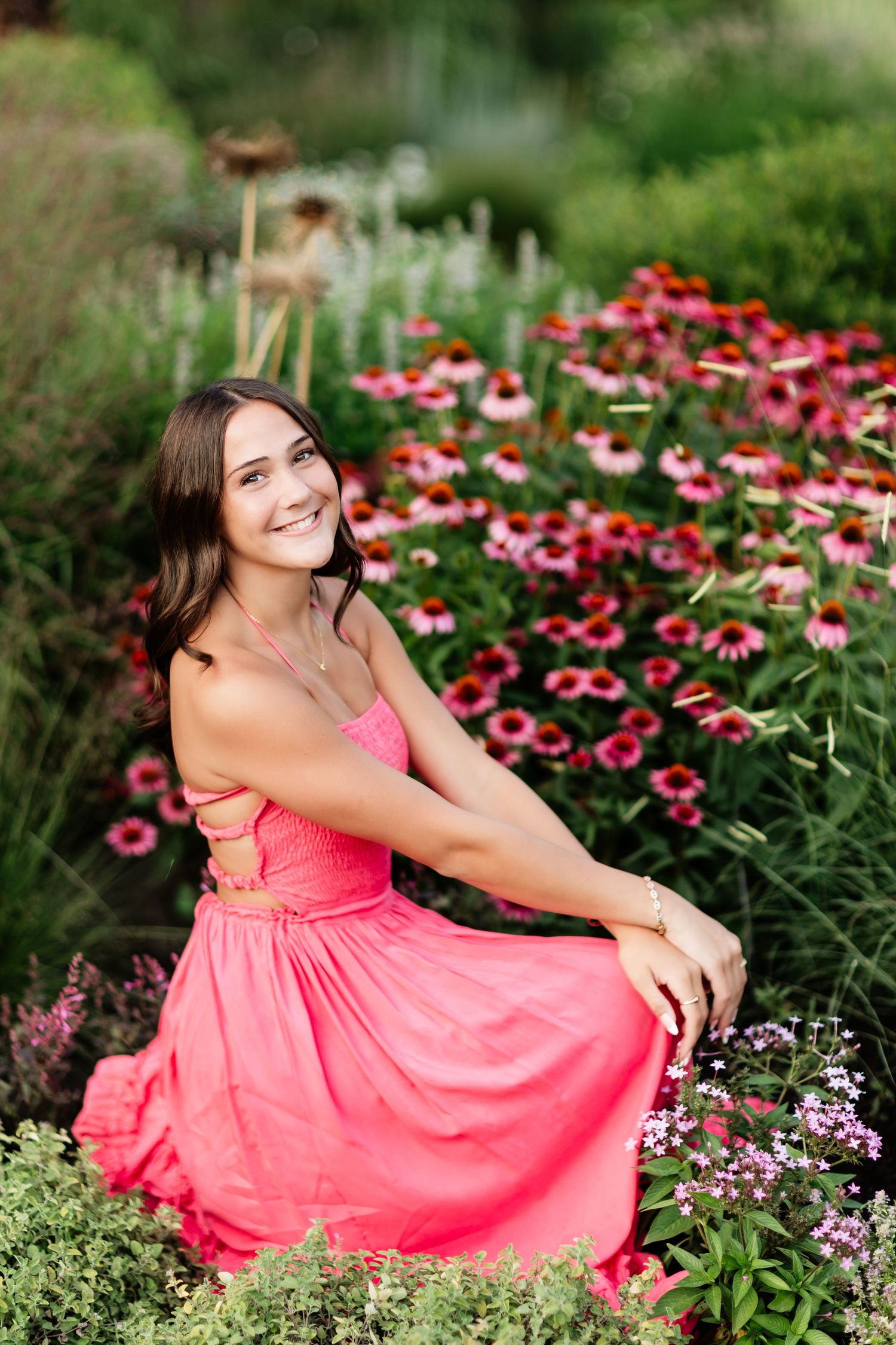 Smiling young woman in a pink maxi dress sitting gracefully among colorful coneflowers in a vibrant garden.