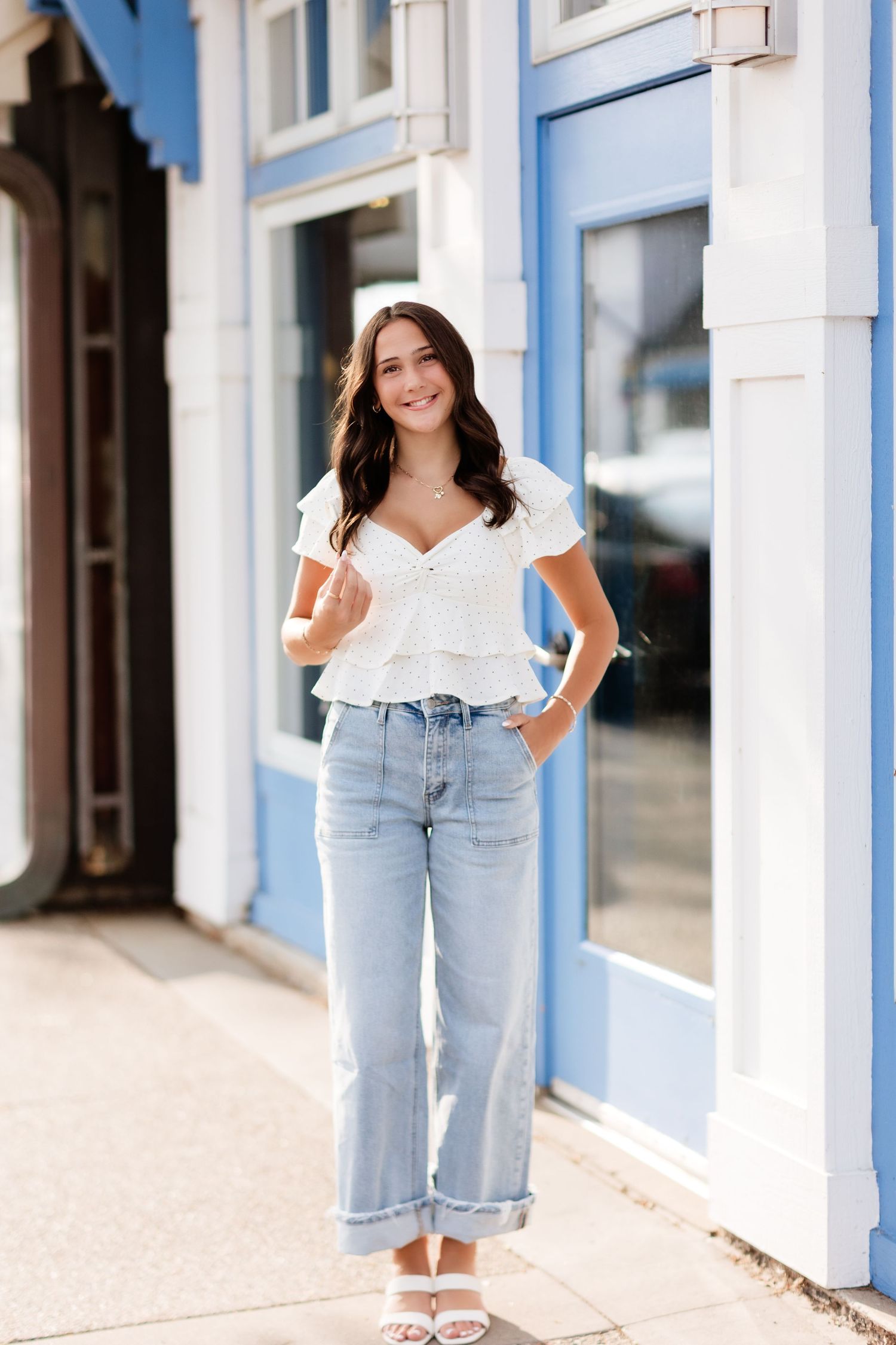 Young woman in white ruffled top and light wash wide-leg jeans posing on sidewalk near blue building.