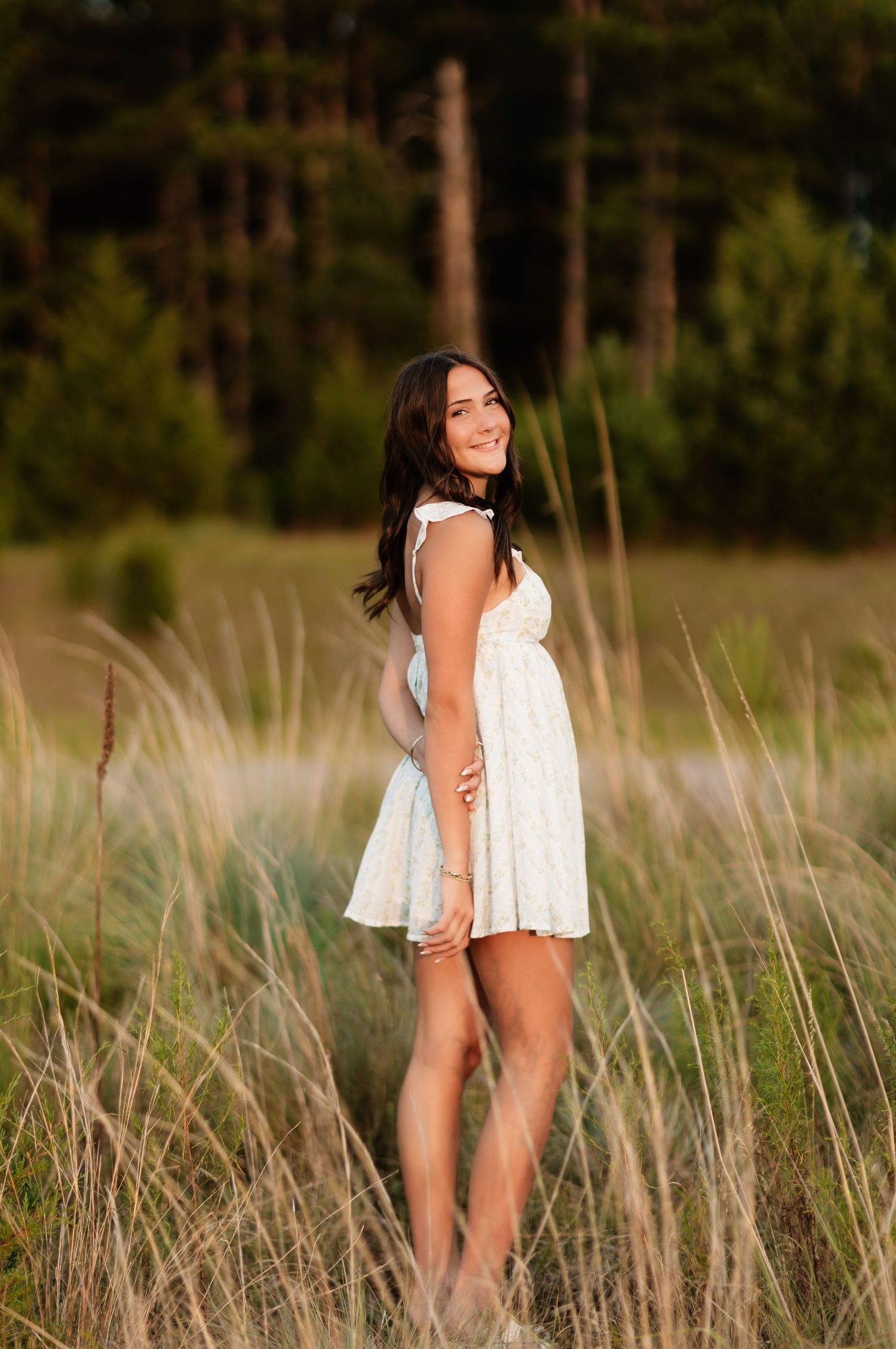 Young woman in white mini dress smiling over her shoulder in golden tall grass near pine trees.