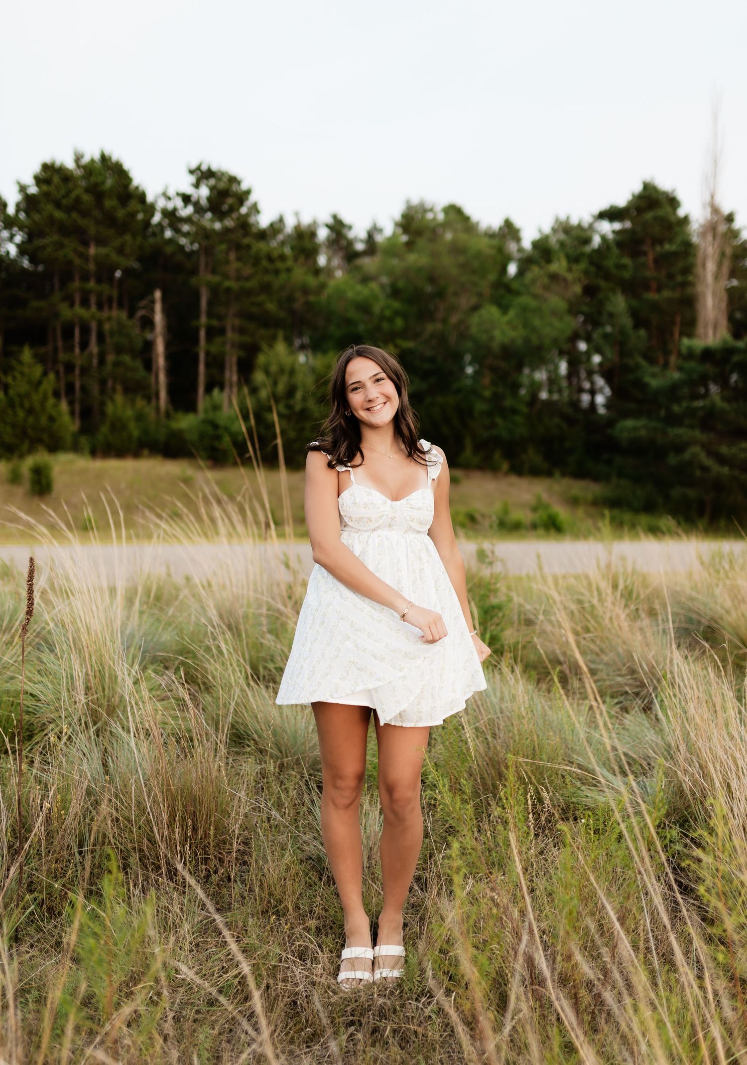Smiling young woman in white floral sundress standing in tall grass with pine trees behind her.