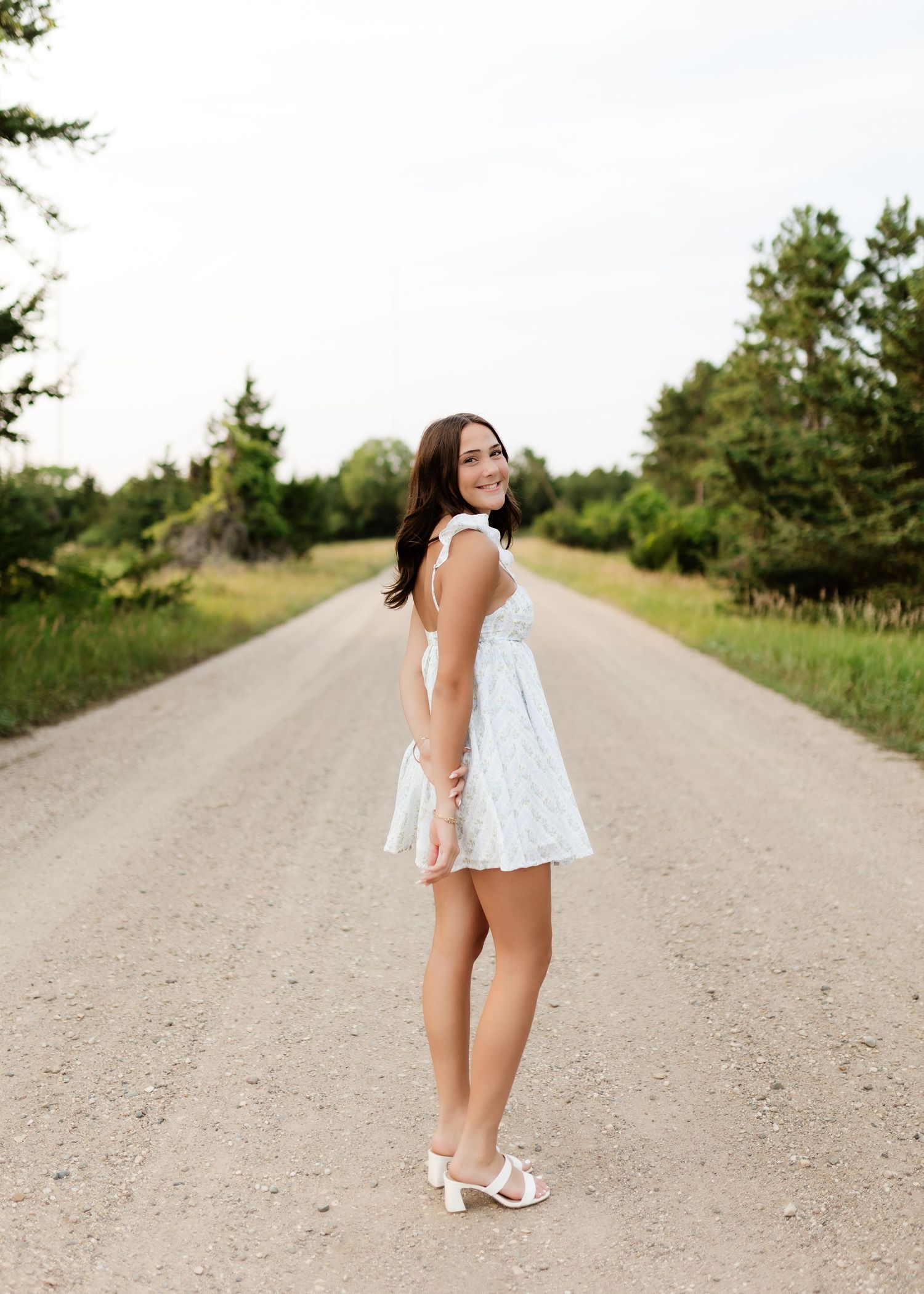 Brunette woman in white mini dress standing on a rural dirt road lined with trees, smiling back.