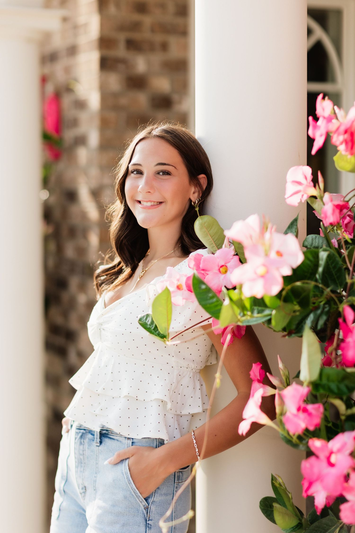 Young woman in white ruffled top smiling beside pink blooming flowers near white pillars.