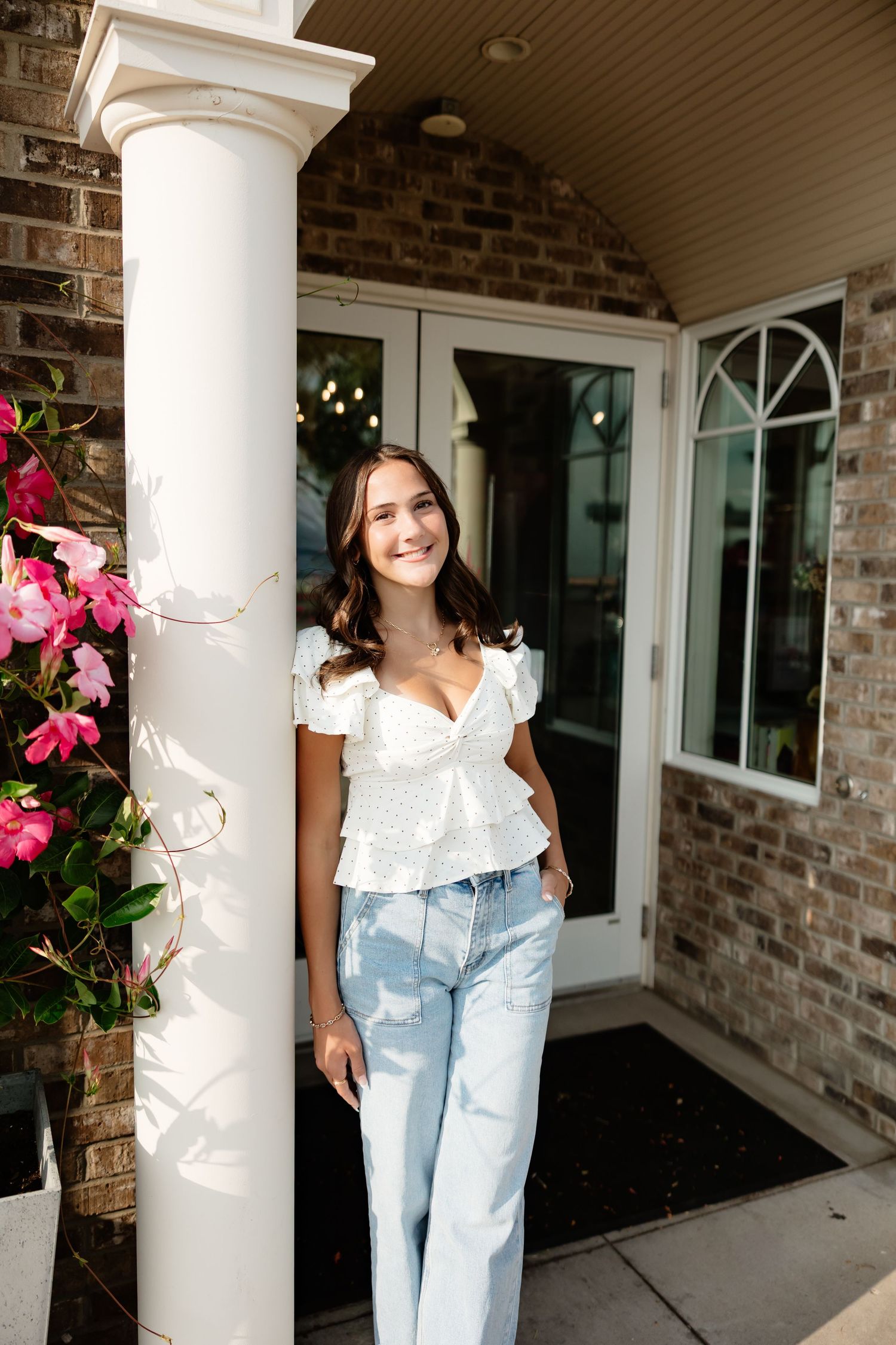 Young woman in white eyelet top and jeans leans against a white column near pink flowers at a brick building.