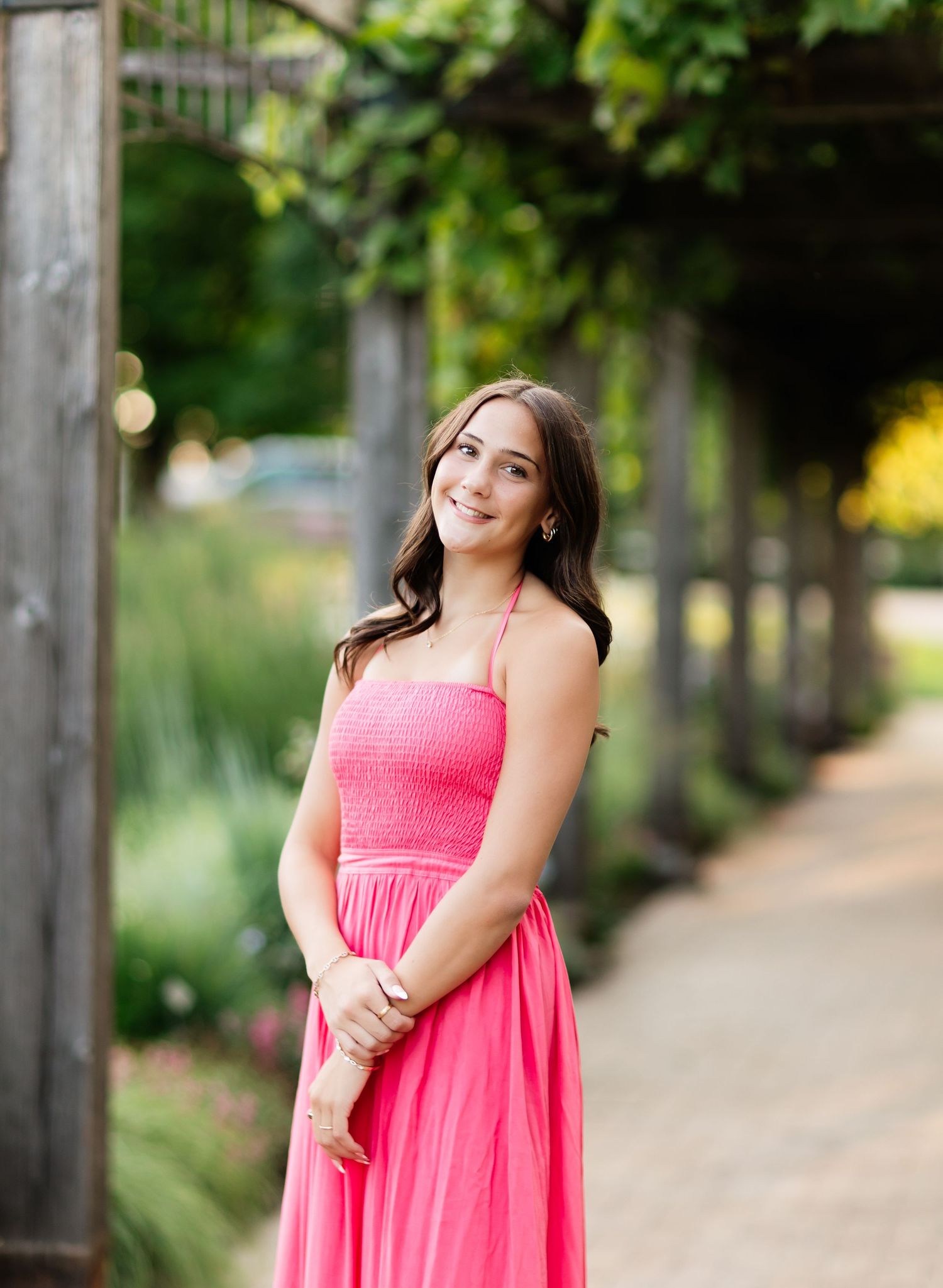 Young woman in pink maxi dress standing on garden pathway under leafy pergola, smiling softly.
