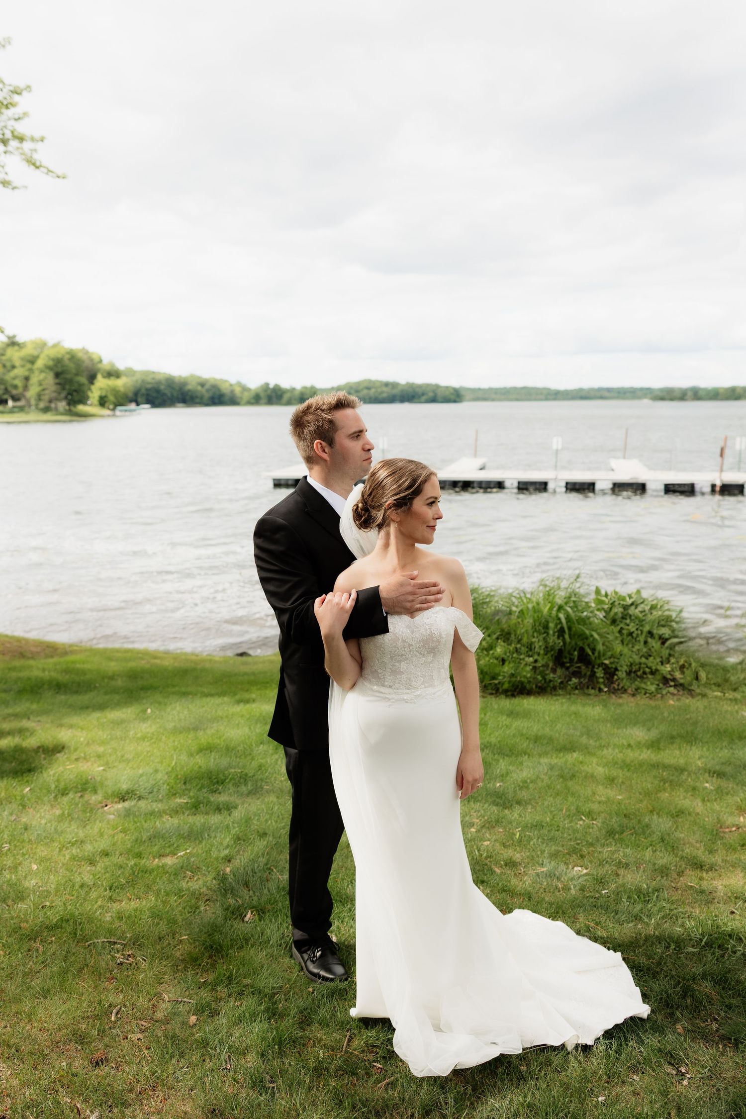 Bride and groom embrace by a lakeside on their wedding day, she in a white gown, he in a black tuxedo.